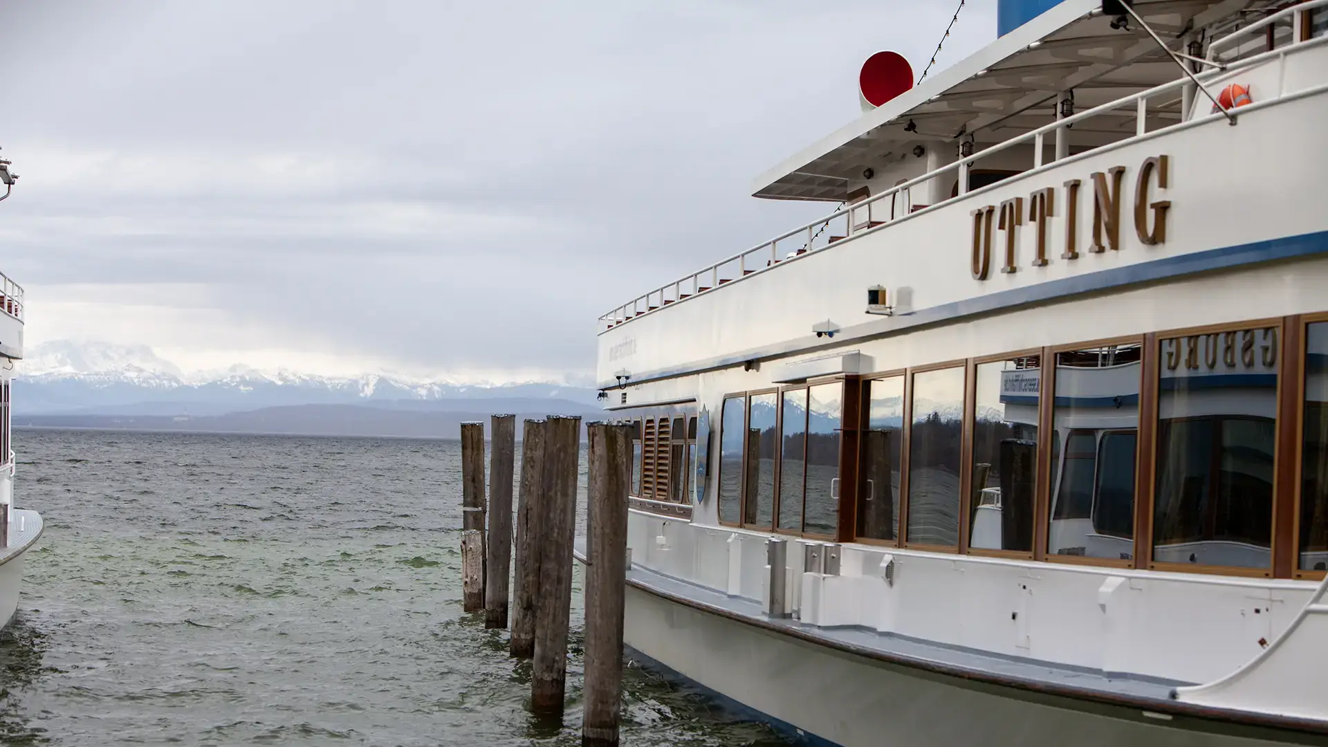 bayerische Seenschifffahrt ammersee - im Heimathafen Stegen - das Schiff MS Utting liegt am Landungssteg und die Sicht auf die Aussenseite mit Schriftzug ist erkennbar. Dahinter der Ammersee und die Silhouette der Berge
