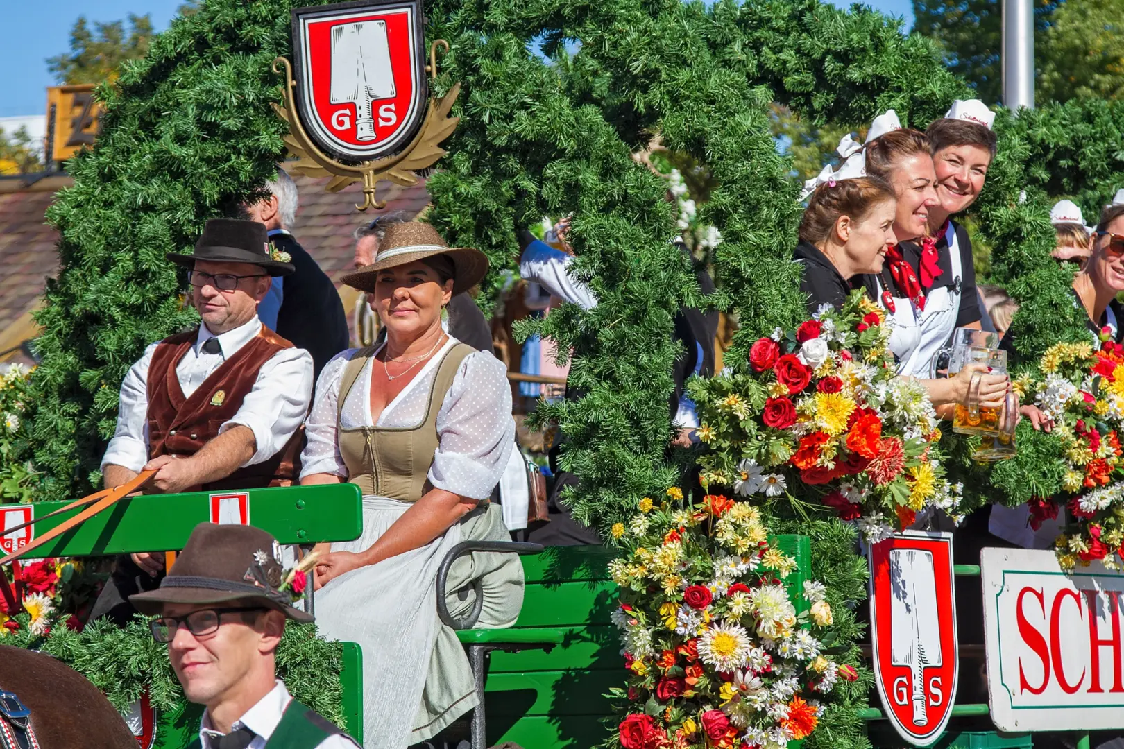 Wirtsleute in Tracht sitzen in einer mit Girlanden und Blumen geschmückten Festkutsche des Schottenhamel-Festzelts, dahinter Bedienungen mit Maßkrügen, Einzug der Festwirte auf dem Oktoberfest München