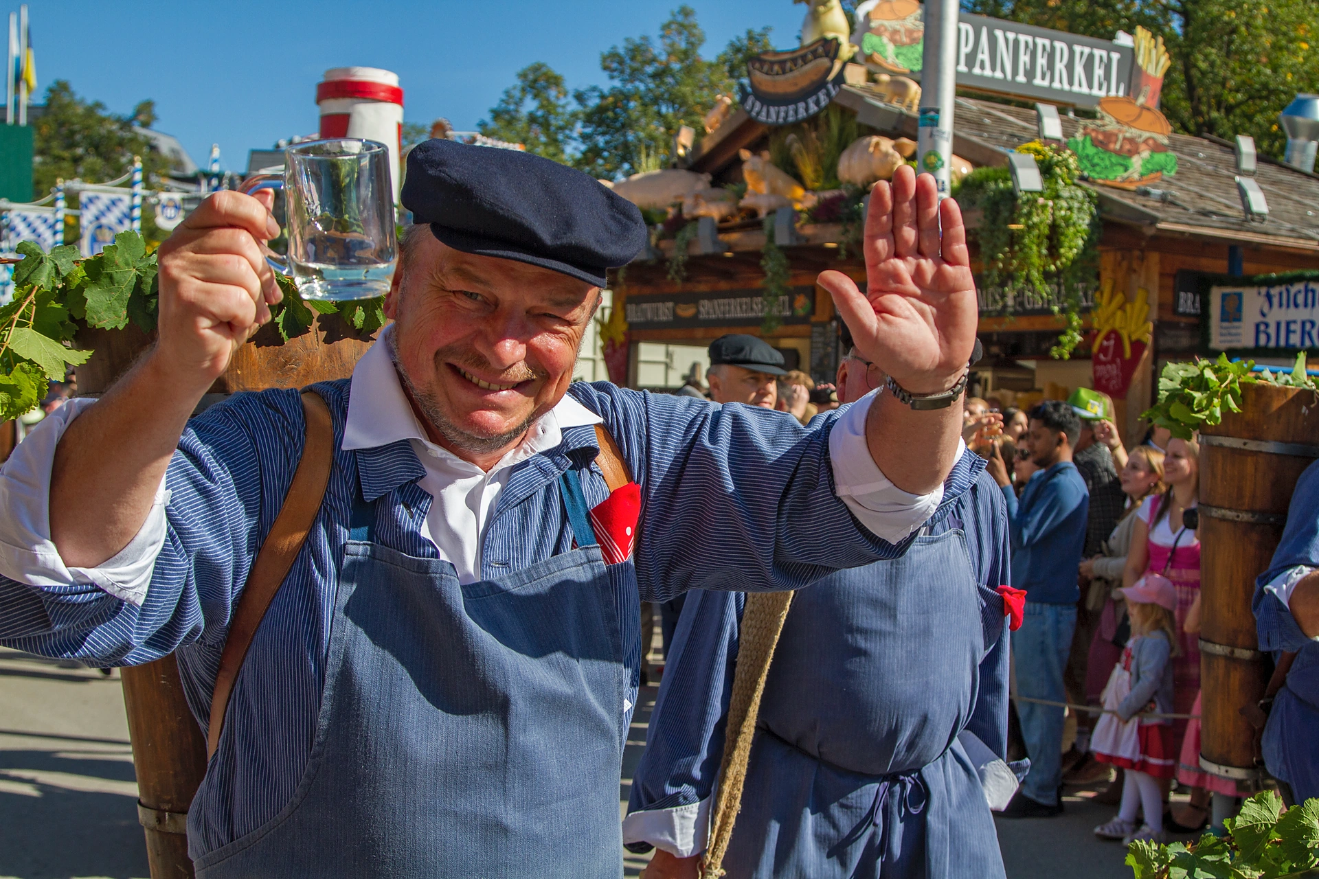 Ein Mann in blauer Arbeitstracht mit Schirmmütze hebt einen Glaskrug und winkt, im Hintergrund der Wagen des Weinzelts, Einzug der Festwirte auf dem Oktoberfest München