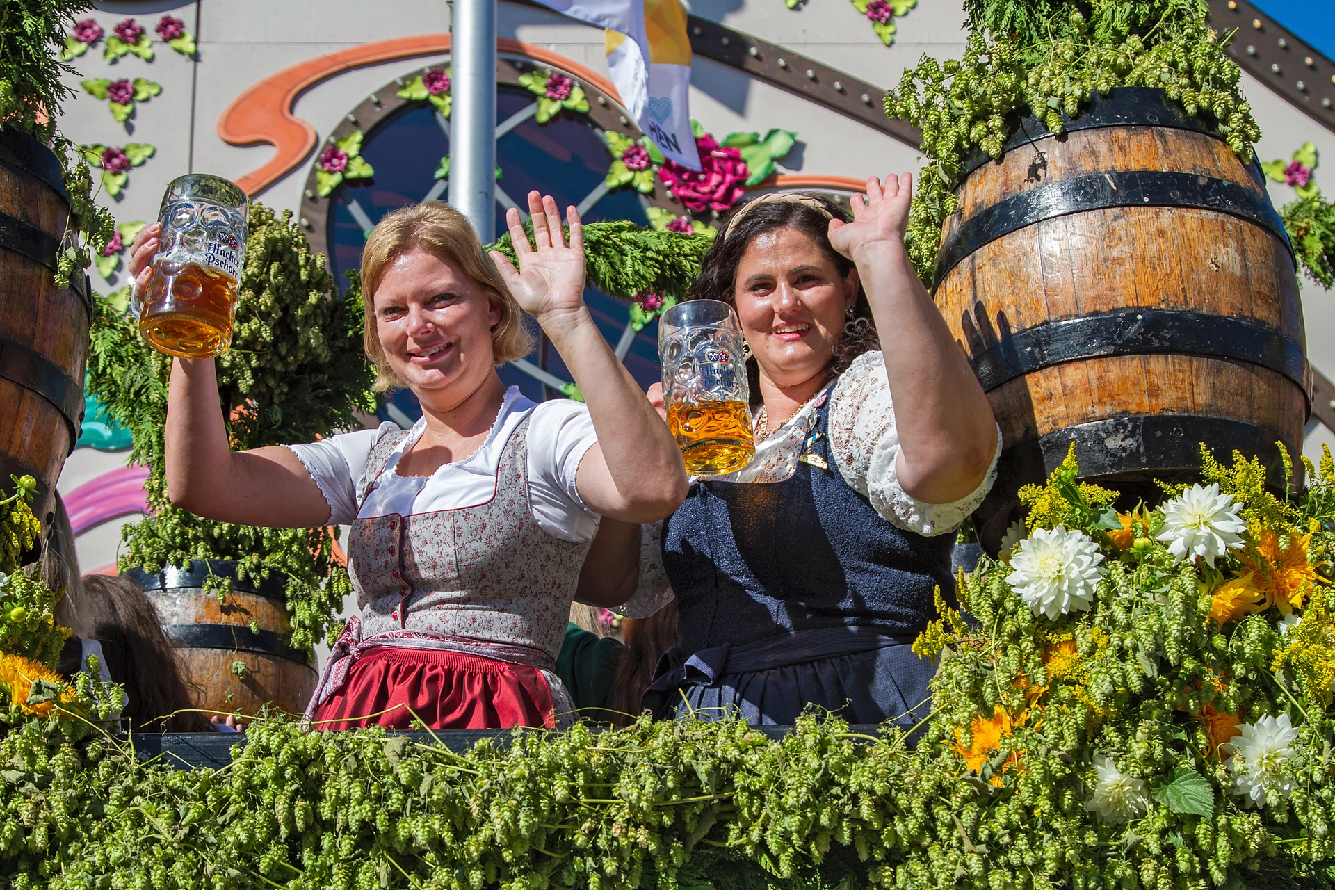 Zwei Frauen in Dirndln heben Maßkrüge und winken von einem Festwagen mit Holzfass und Hopfenranken, Einzug der Festwirte auf dem Oktoberfest München