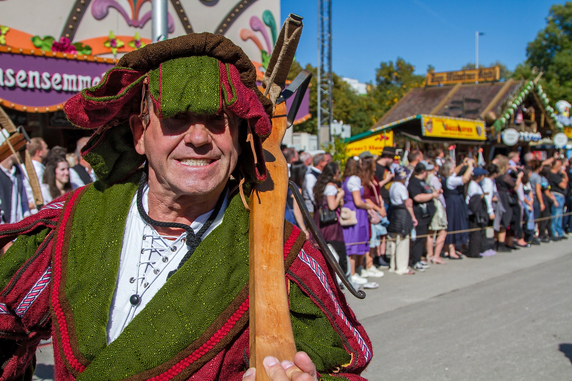 Ein Mann in historischer Tracht mit moosgrünem Umhang und Hut trägt eine Holzstange über der Schulter und lacht in die Kamera, Einzug der Festwirte auf dem Oktoberfest München