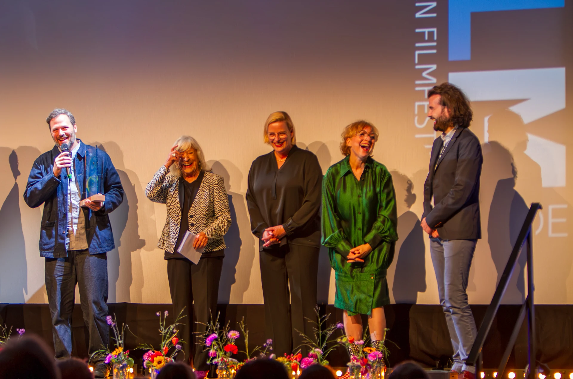 Sebastian Brameshuber spricht mit Trophäe und Mikrofon auf der Bühne der Schlossberghalle Starnberg, daneben Margarethe von Trotta und weitere Jurymitglieder
