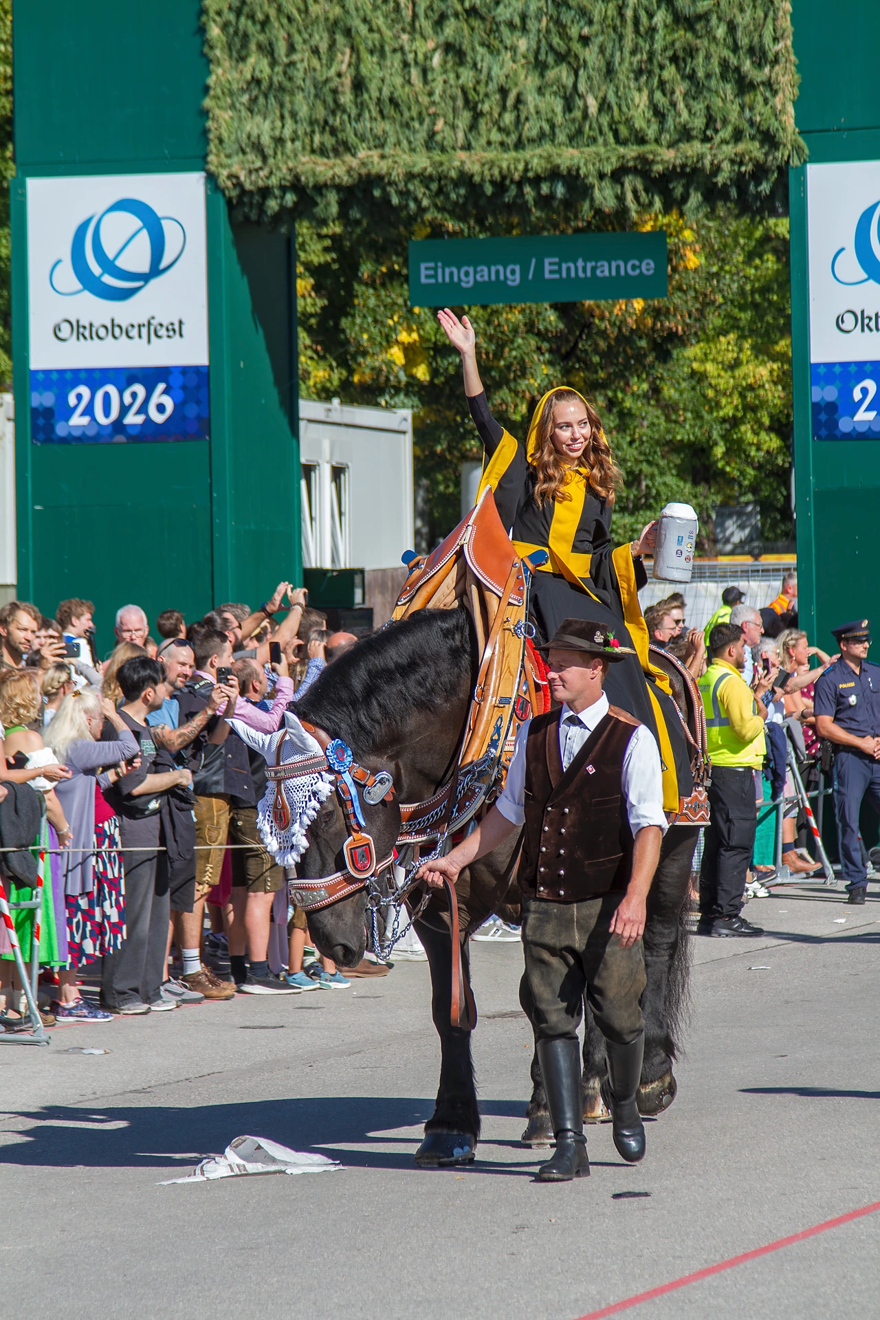 Das Münchner Kindl Sina Hochreiter winkt im schwarz-gelben Ornat vom Kaltblut, in der linken Hand einen Maßkrug, daneben führt ein Mann in Tracht das Pferd, im Hintergrund das Oktoberfest-Banner 2026 am Eingang zur Theresienwiese