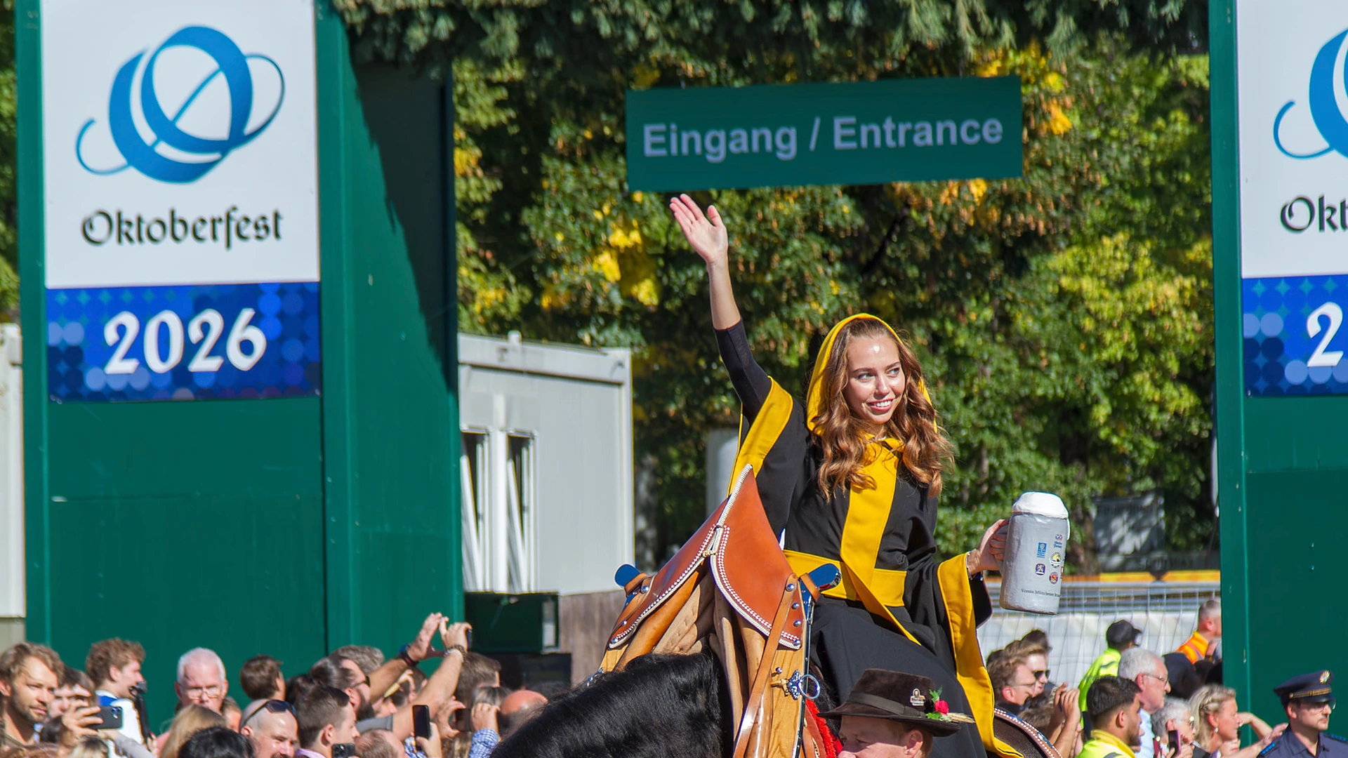 fuenfseen-muenchner-kindl-oktoberfest-20260919-c-bwidmann-5153-titel-1920×1080 Das Münchner Kindl Sina Hochreiter winkt im schwarz-gelben Ornat vom Kaltblut, in der linken Hand einen Maßkrug, daneben führt ein Mann in Tracht das Pferd, im Hintergrund das Oktoberfest-Banner 2026 am Eingang zur Theresienwiese