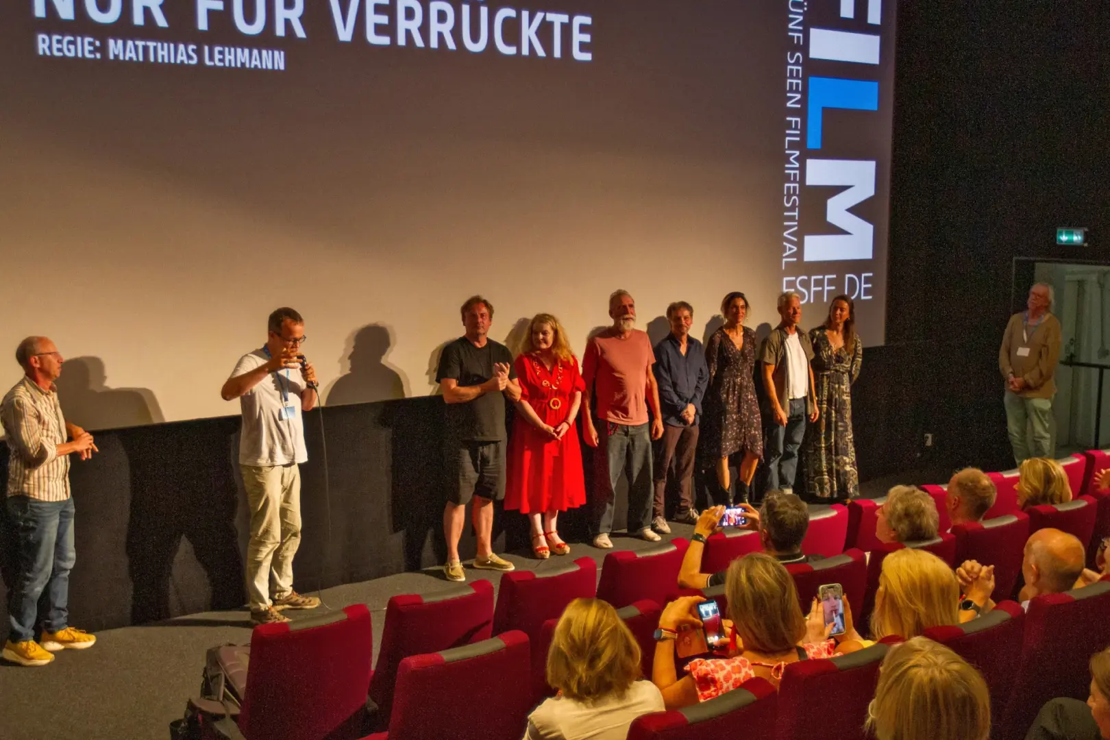 Regisseur Matthias Lehmann spricht ins Mikrofon, dahinter das Filmteam von Nur für Verrückte auf der Bühne des Breitwand Kinos in Starnberg