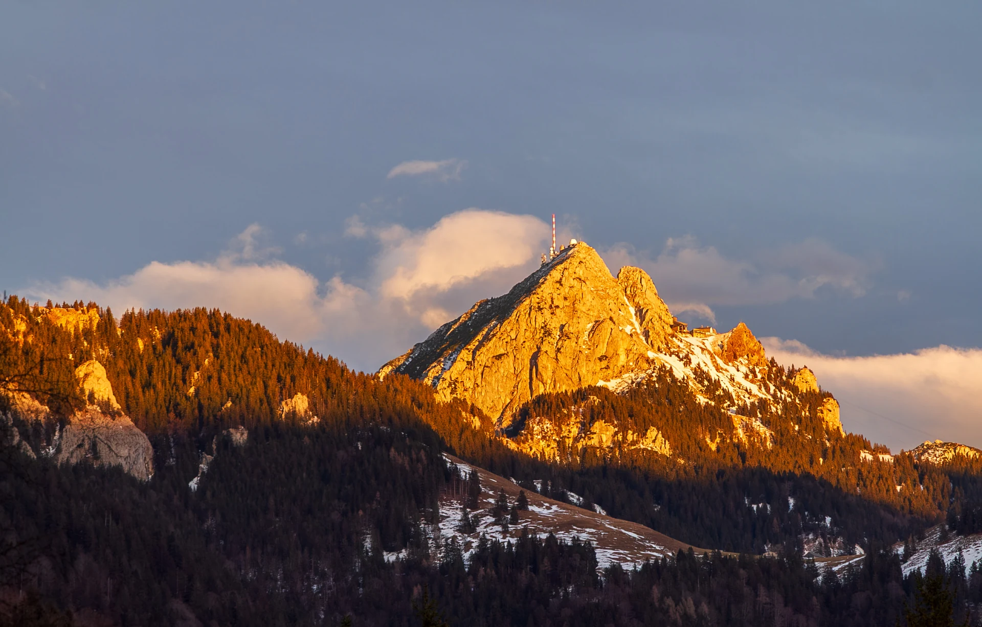 Felsgipfel mit Sendemast im goldenen Abendlicht, ringsum verschneite Wälder und Wolken