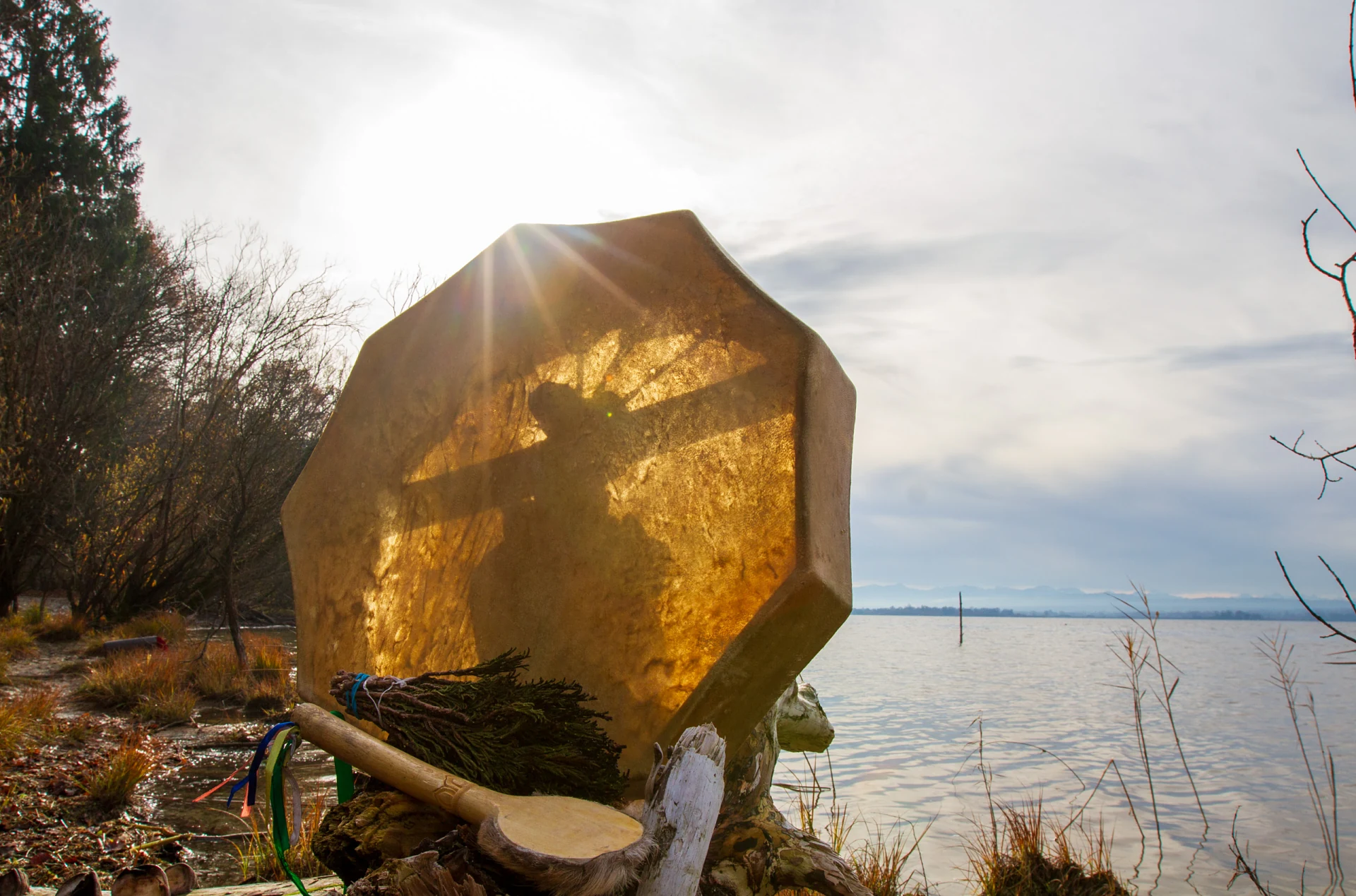 Achteckige Rahmentrommel im Gegenlicht am Ufer des Ammersees, die Sonne strahlt über den oberen Rand, davor ein Wacholderbüschel und bunte Bänder