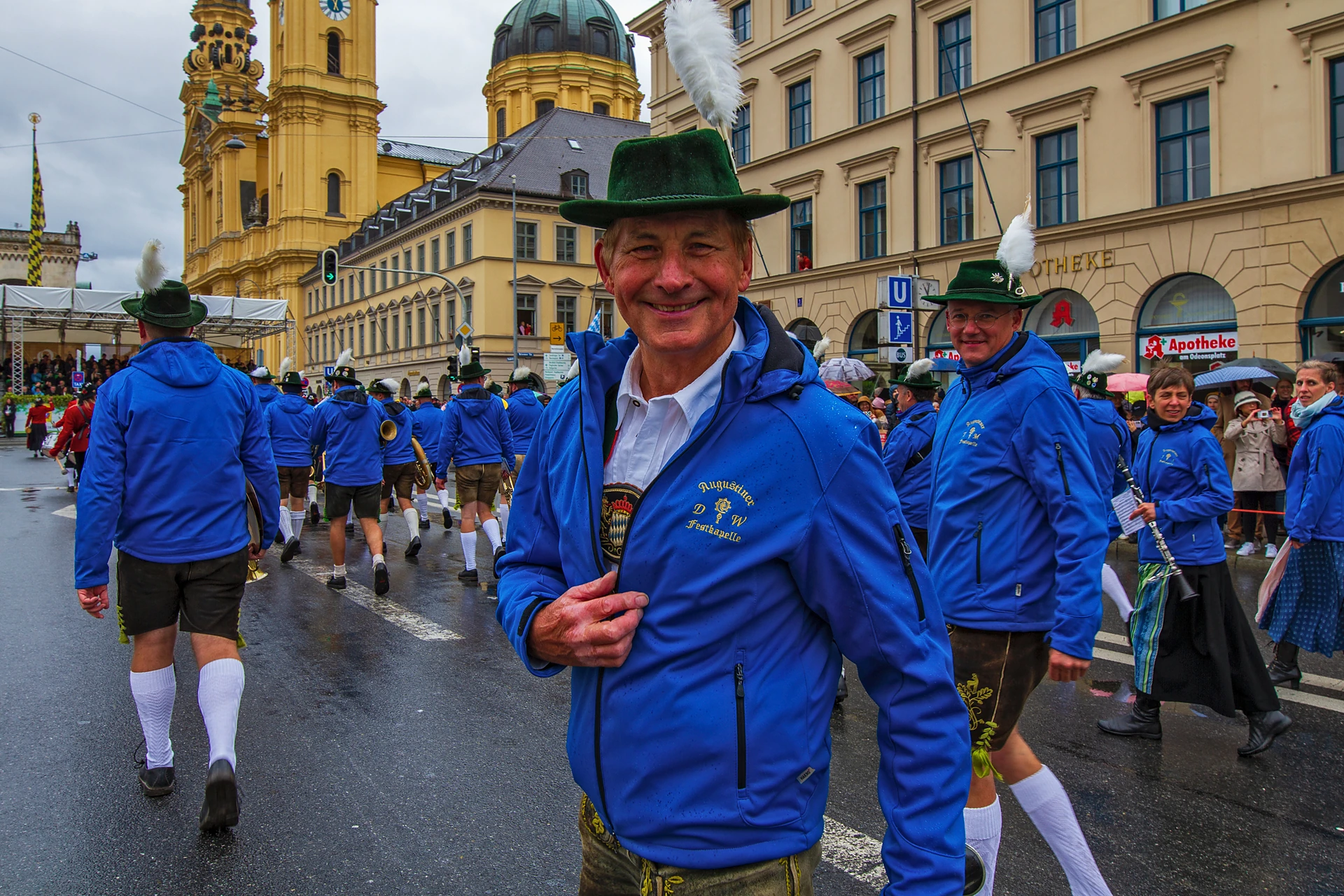 Musiker der Augustiner Festkapelle in blauen Jacken und Trachtenhüten ziehen im Regen am Odeonsplatz vorbei, im Hintergrund die Theatinerkirche