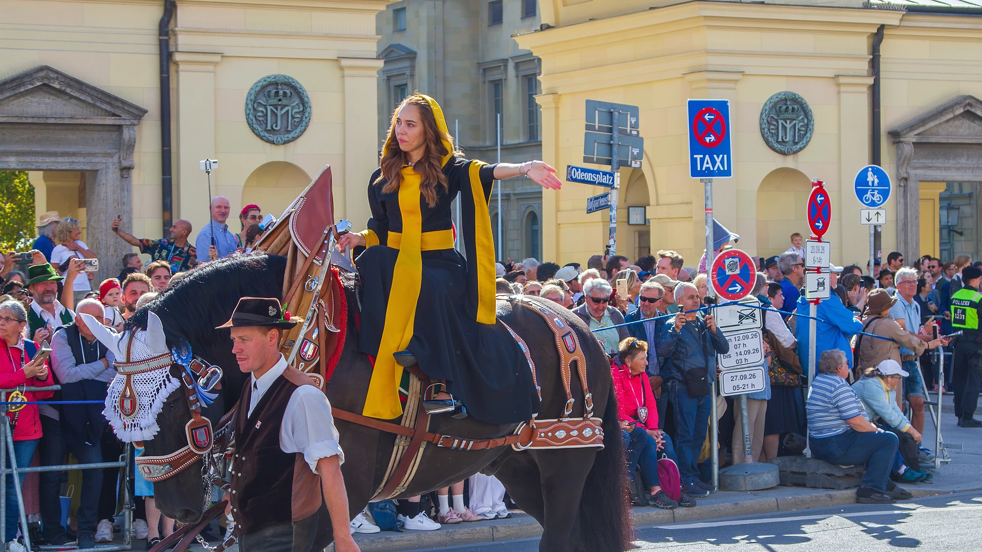 fuenfseen-trachtenzug-oktoberfest-20260920-c-bwidmann-6433-titel Das Münchner Kindl Sina Hochreiter in schwarz-gelber Kutte winkt vom Kaltblutpferd, ein Mann in Tracht führt das Pferd am Zügel, am Straßenrand dicht gedrängte Zuschauer, Trachten- und Schützenzug München