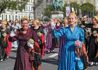 Frauen in historischen Kleidern mit goldbestickten Hauben und Blumensträußen winken den Zuschauern, Trachten- und Schützenzug München