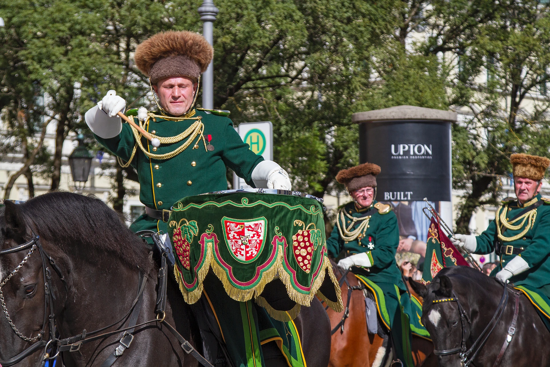 Ein Reiter in grüner Uniform mit Pelzmütze hebt grüßend die Hand, sein Pferd trägt eine bestickte Satteldecke, Trachten- und Schützenzug München