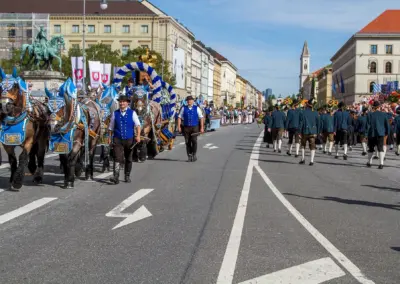 Ein Brauereigespann mit blau-weißem Schmuck zieht einen Festwagen durch die Ludwigstraße, dahinter eine Kapelle in grauen Uniformen, Trachten- und Schützenzug München