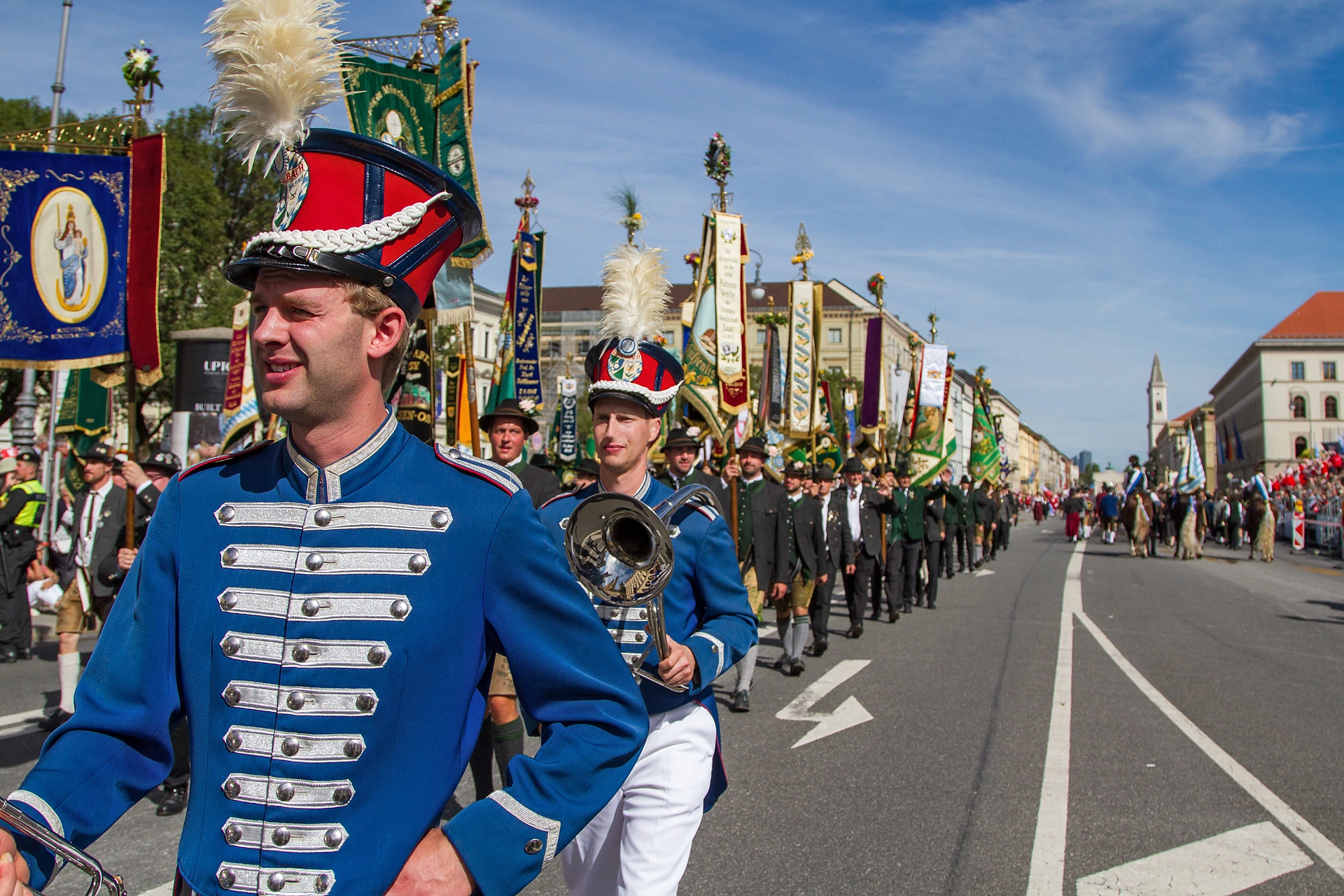 Ein junger Musiker in blauer Uniform mit weißen Schnüren und rotem Tschako lacht in die Kamera, hinter ihm sein Spielmannszug, Trachten- und Schützenzug München