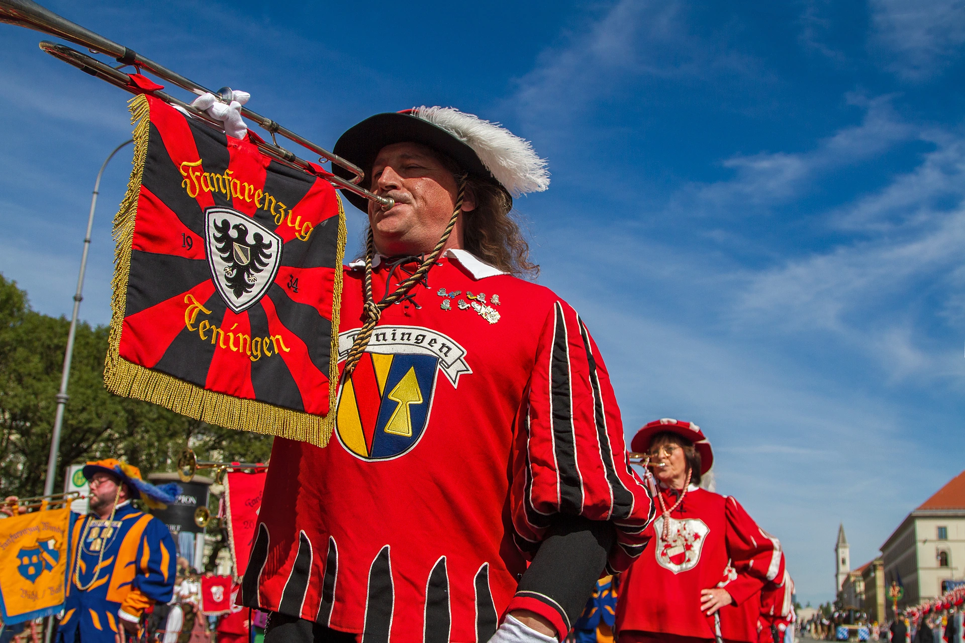 Ein Fanfarenbläser in rot-gelbem Wams mit Federhut hält eine rote Fahne mit Wappen vor blauem Himmel, Trachten- und Schützenzug München