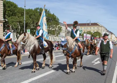 Reiter in Tracht aus verschiedenen Vereinen von Pferdeland Bayern auf Pferden mit Fahnen ziehen durch die Ludwigstraße, einer winkt mit dem Hut, Trachten- und Schützenzug München