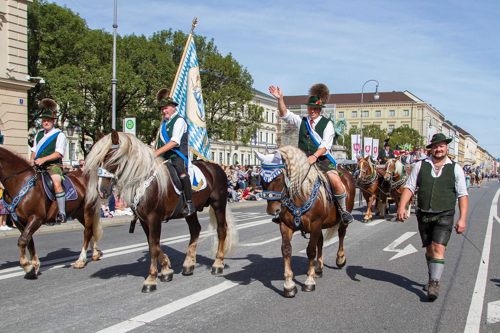 Reiter in Tracht aus verschiedenen Vereinen von Pferdeland Bayern auf Pferden mit Fahnen ziehen durch die Ludwigstraße, einer winkt mit dem Hut, Trachten- und Schützenzug München