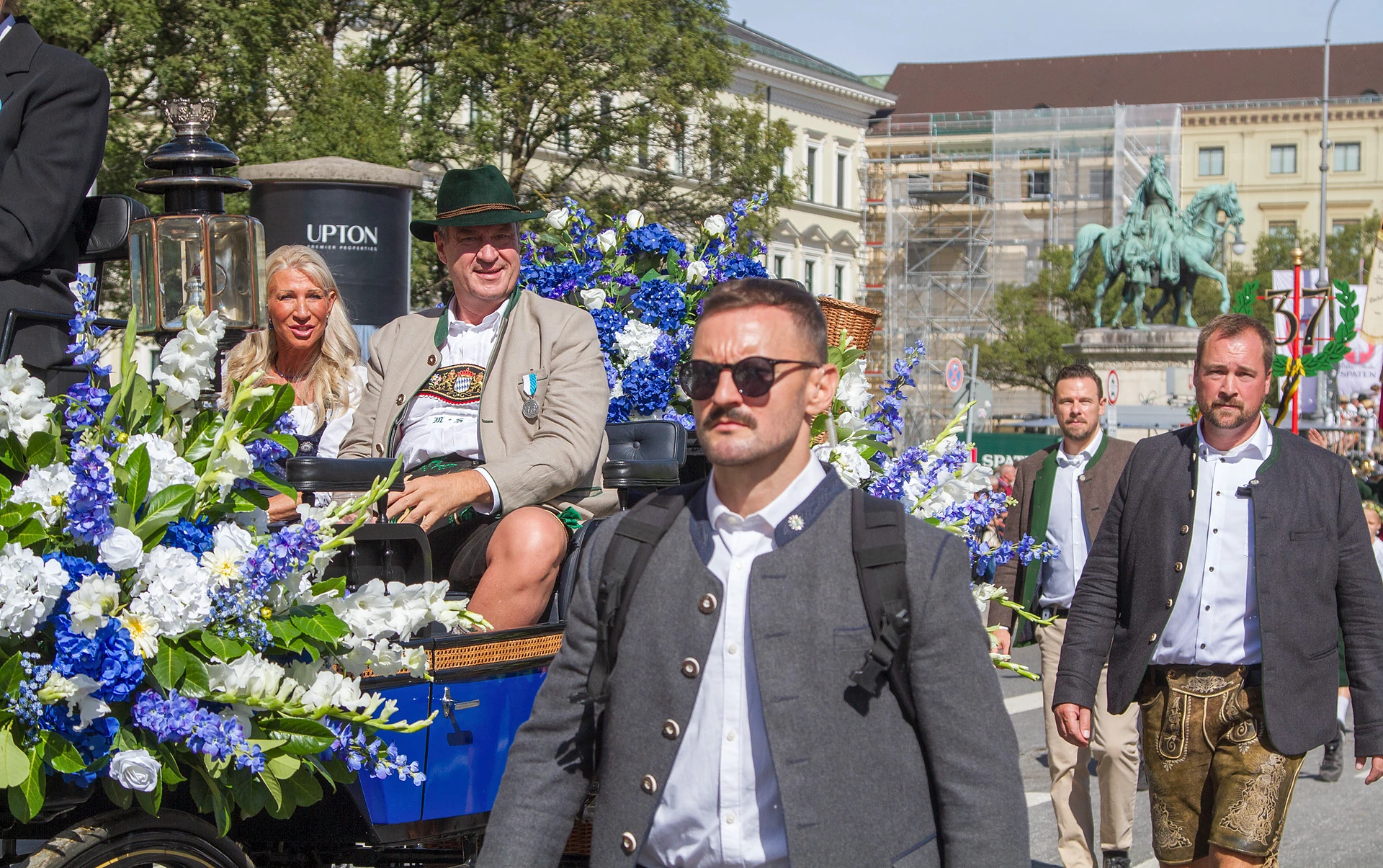 Ministerpräsident Markus Söder und seine Frau sitzen in einer mit weißen und blauen Blumen geschmückten Festkutsche, davor Begleitpersonen, Trachten- und Schützenzug München
