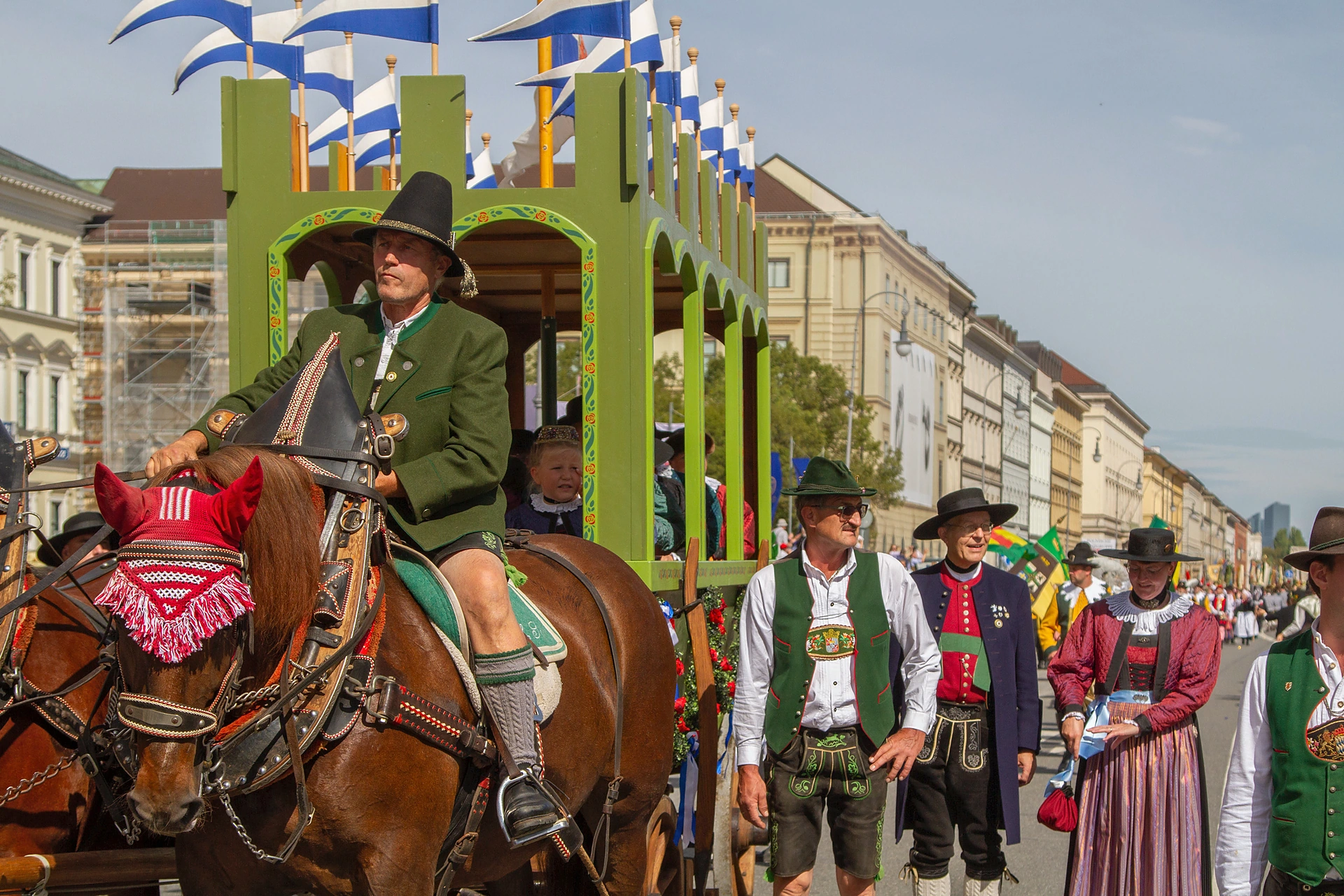 Ein Reiter in grüner Tracht führt einen grünen, blau-weiß geschmückten Festwagen, daneben Trachtlerinnen und Trachtler, Trachten- und Schützenzug München