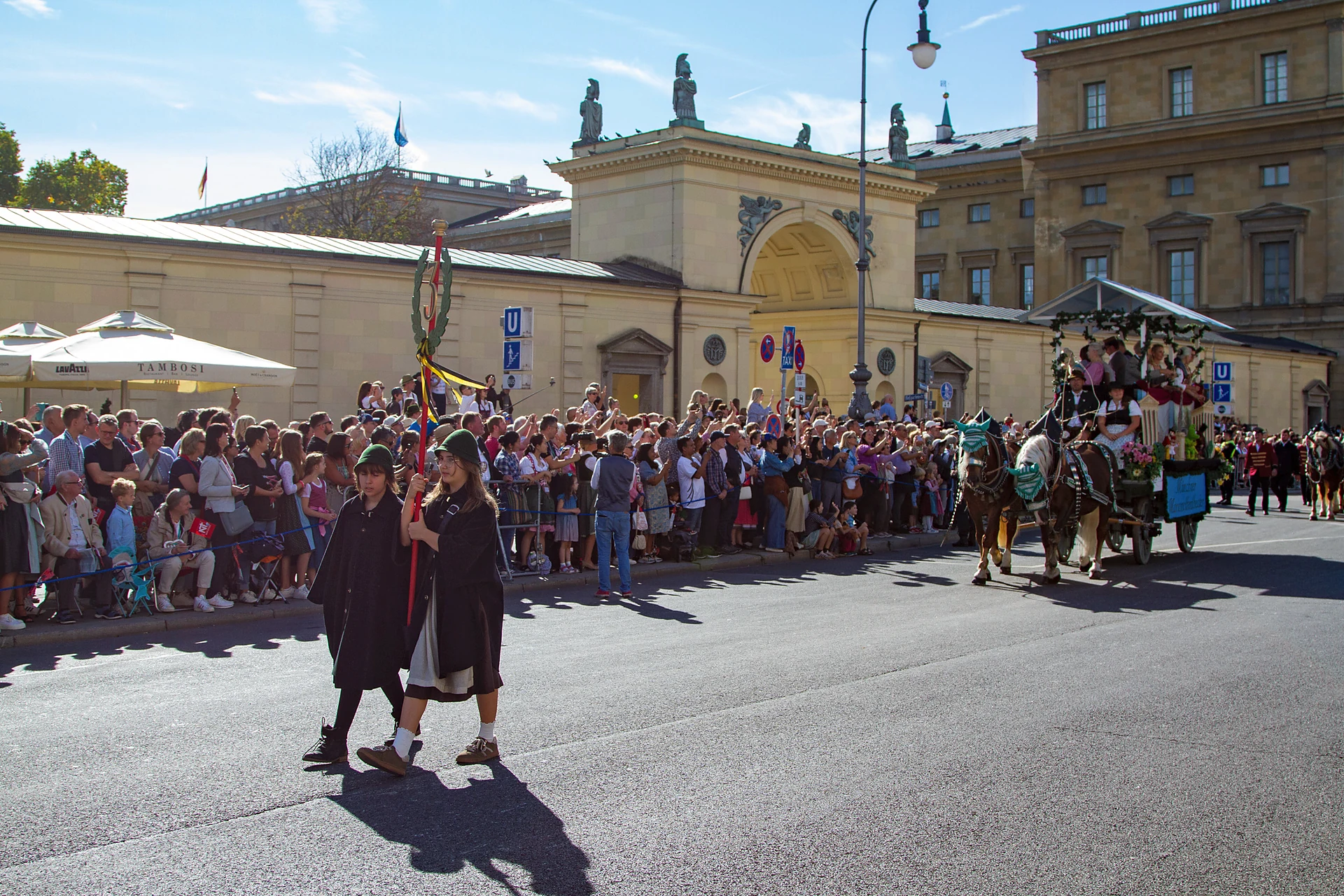 Zwei Taferlträger in dunklen Capes gehen mit dem Zugschild vor ihrer Gruppe, im Hintergrund Zuschauer und eine Kutsche, Trachten- und Schützenzug München