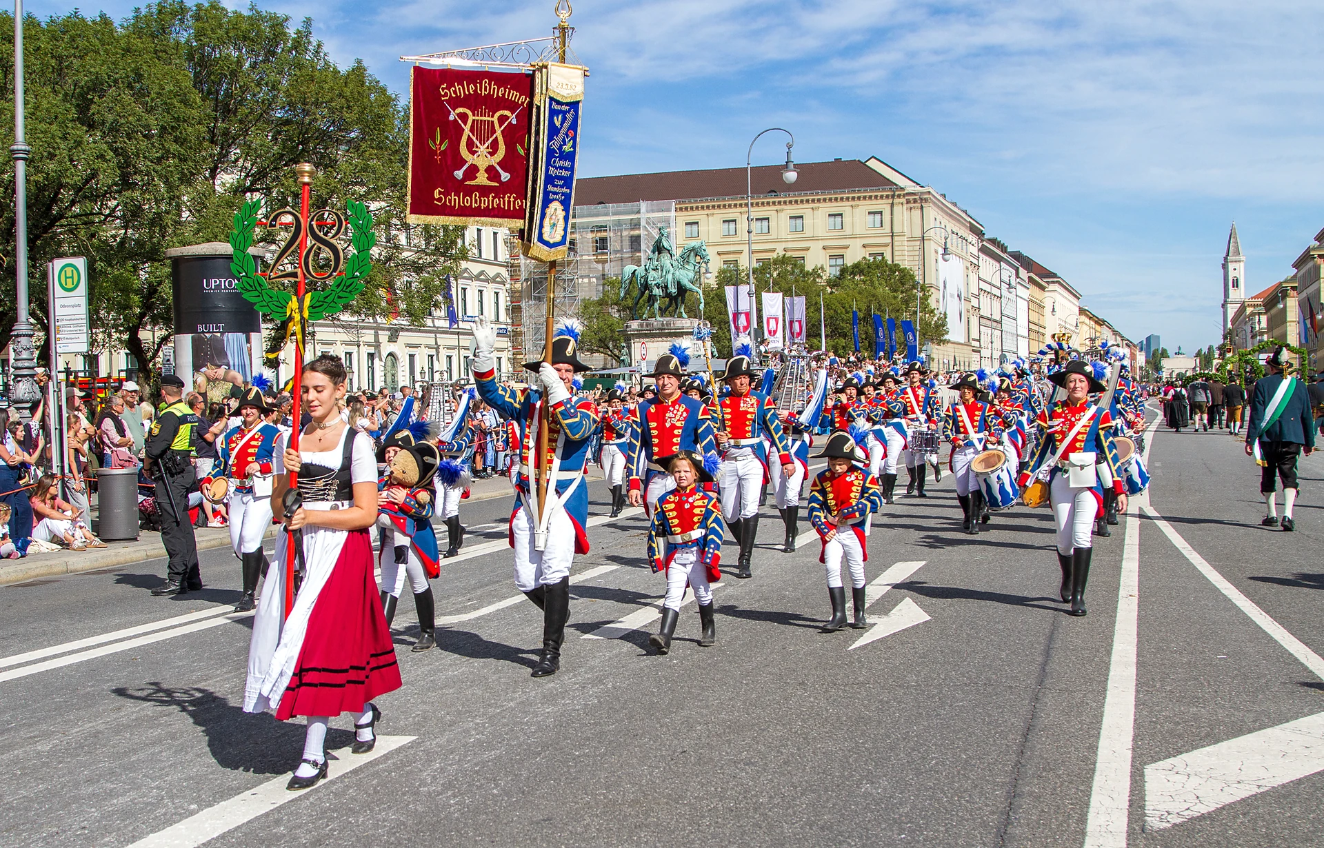 Eine junge Frau im Dirndl geht vor dem Zugschild und einer Kapelle in blau-roten Uniformen, Trachten- und Schützenzug München