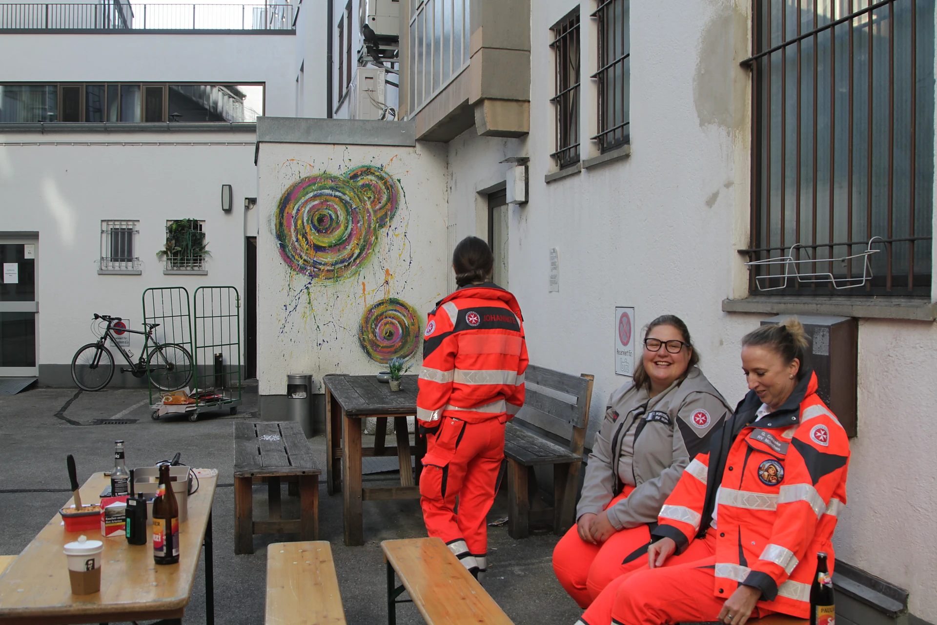 Drei Einsatzkräfte der Johanniter in roter und grauer Dienstkleidung in einem Innenhof, zwei von ihnen sitzen lachend auf einer Bank, München