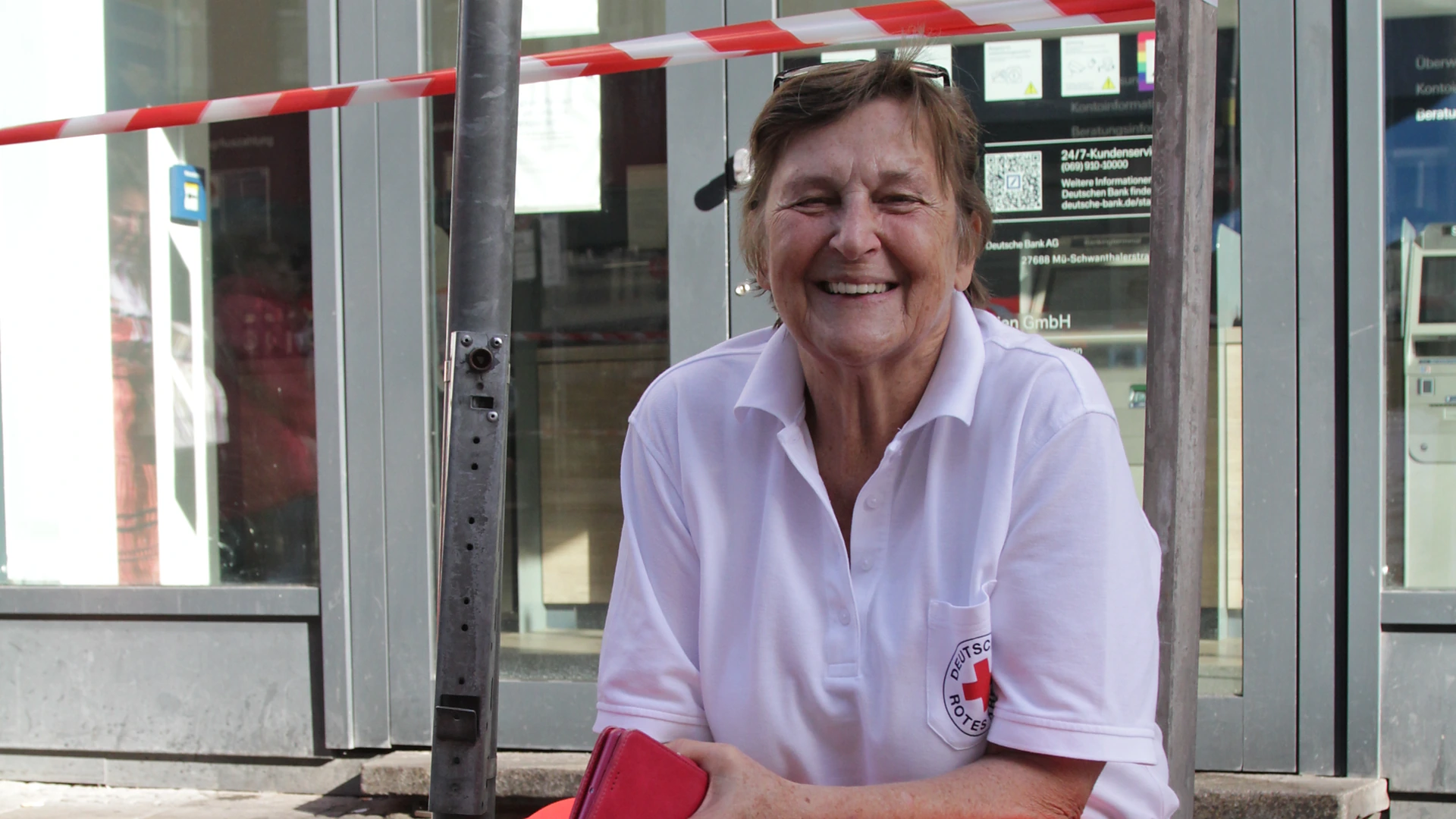 fuenfseen-wiesn-ehrenamt-20260920-c-bwidmann-6052-titel Die ehrenamtliche Rotkreuz-Sanitäterin Irmgard sitzt lachend auf Absperrsockeln am Straßenrand, über ihr rot-weißes Absperrband, München