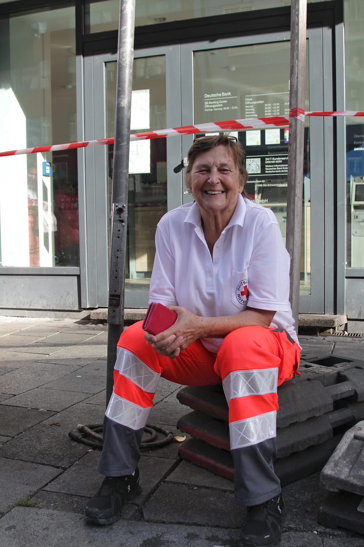 Die ehrenamtliche Rotkreuz-Sanitäterin Irmgard sitzt lachend auf Absperrsockeln am Straßenrand, über ihr rot-weißes Absperrband, München