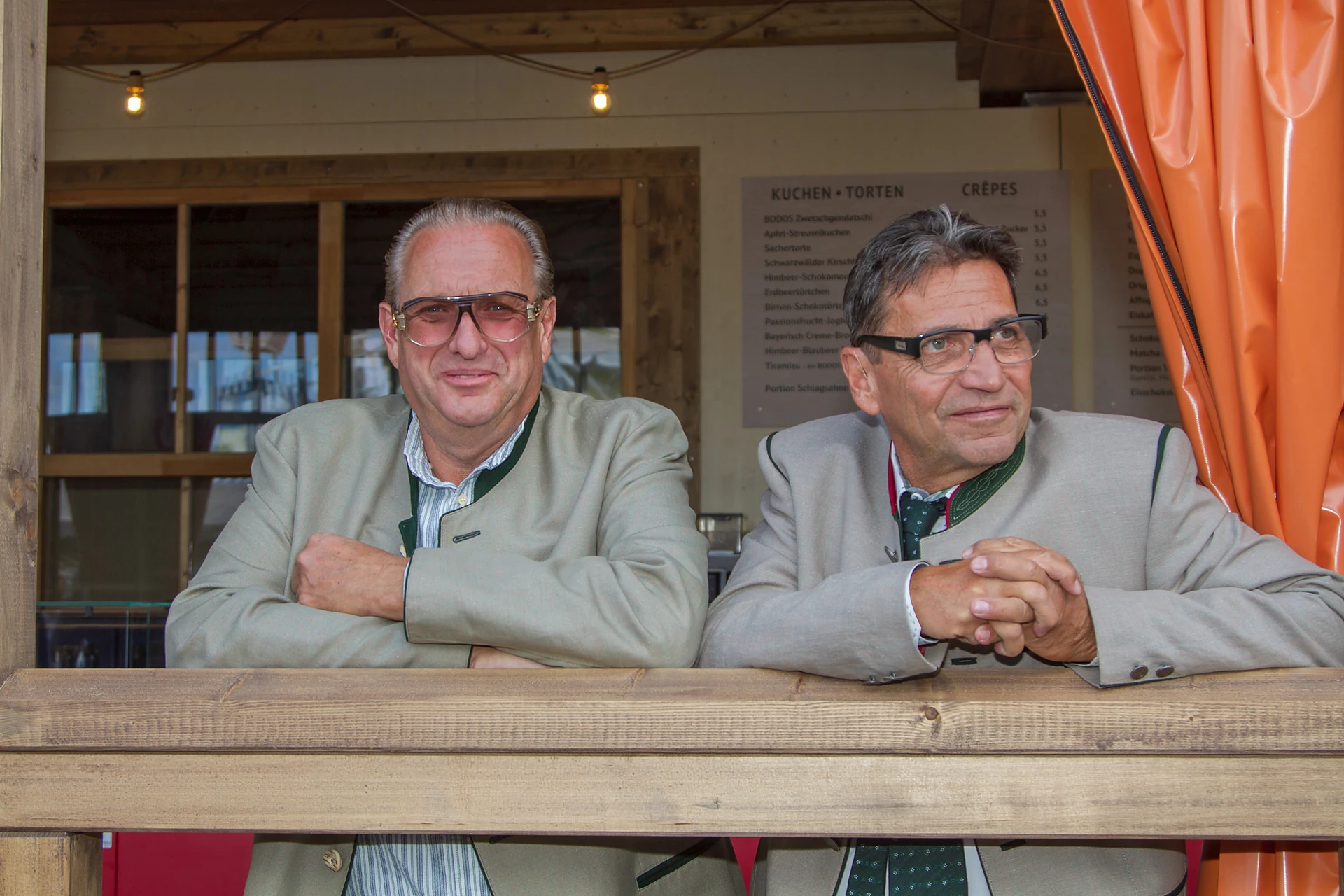 Robert Engel mit getönter Brille und ein zweiter Mann in Trachtenjanker lehnen am offenen Holztresen von Bodos Cafézelt, im Hintergrund die Kuchenkarte, Oktoberfest München PersonInImage: Robert Engel