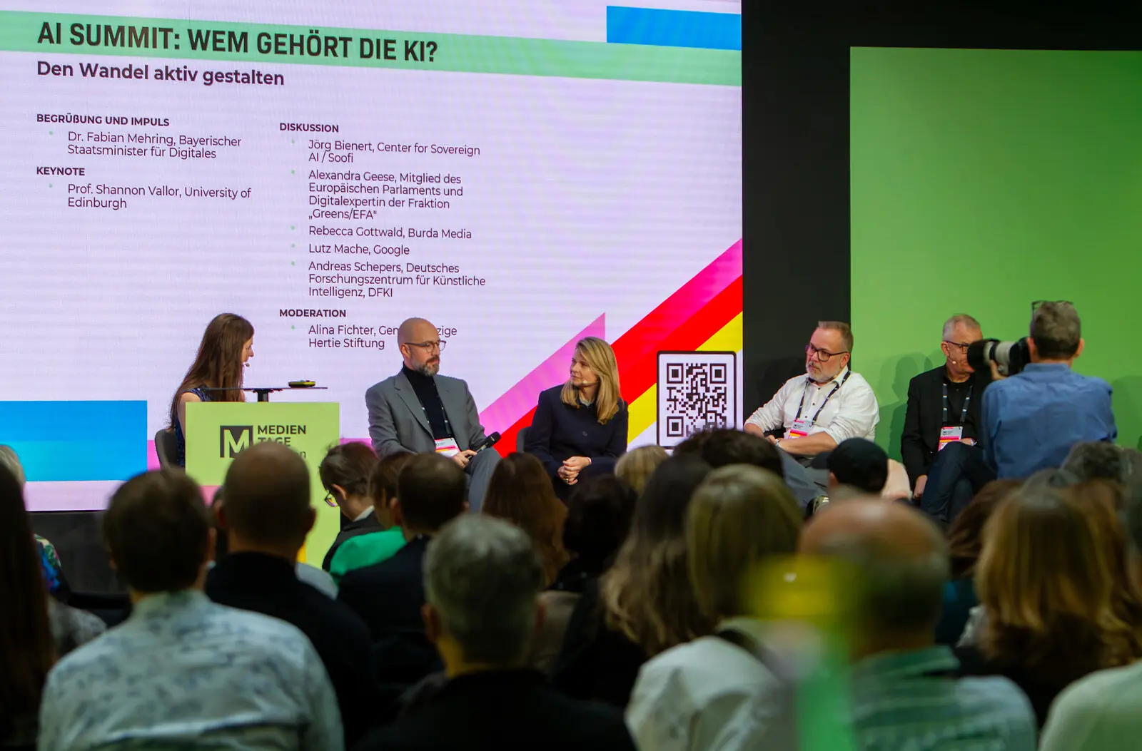 im Hintergrund eine große Präsentation mit viel Text und Namen auf der Bühne sitzend Frauen und Männer diskutierend über das Thema im Vordergrund die Hinterköpfe des Publikums