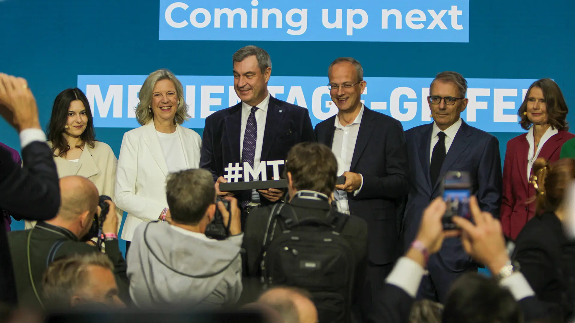 Sechs Personen stehen nebeneinander auf einer Bühne und halten ein Schild mit der Aufschrift Hashtag M T M, davor eine Reihe fotografierender Journalisten.