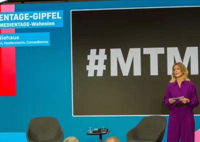 Eine Frau im violetten Anzug steht mit Karten in der Hand auf einer Bühne vor einer Leinwand mit dem Schriftzug Hashtag M T M 40.