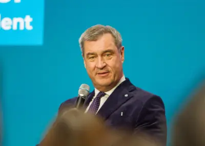 Ein Mann im dunklen Anzug spricht in ein Handmikrofon, im Hintergrund eine blaue Wand mit einer Texteinblendung.