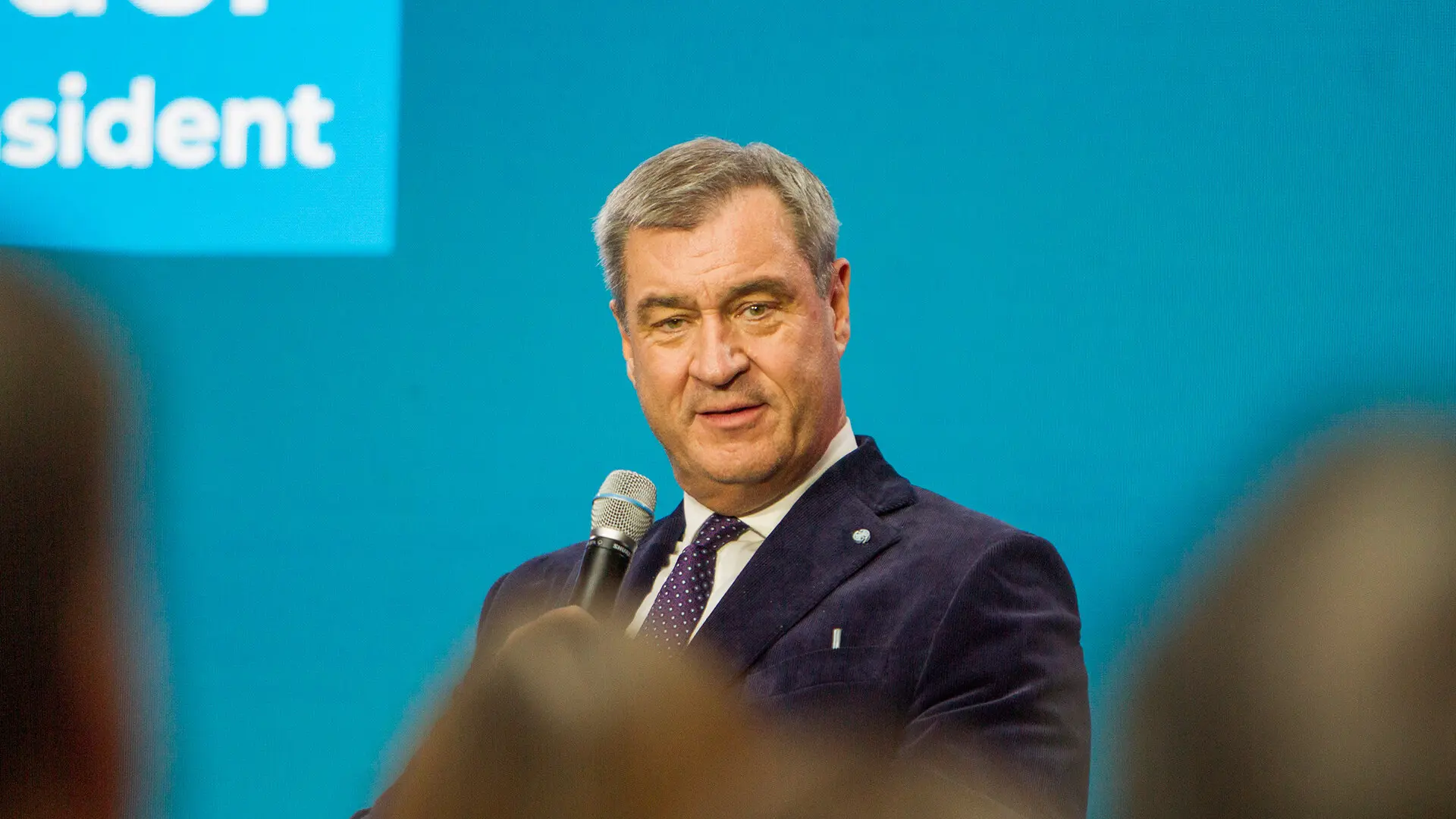 Ein Mann im dunklen Anzug spricht in ein Handmikrofon, im Hintergrund eine blaue Wand mit einer Texteinblendung.