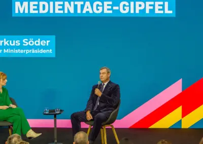 großer weisser Schriftzug Medientage-Gipfel auf blauem Hintergrund links daneben Dr. Markus Söder Bayerischer Ministerpräsident, auf der Bühne sitzend in grünem Kostüm Eva Schulz dazwischen ein Tisch mit zwei Wasser und rechts sitzend Dr. Markus Söder ins Mikrofon sprechend mit dunklem Anzug.
