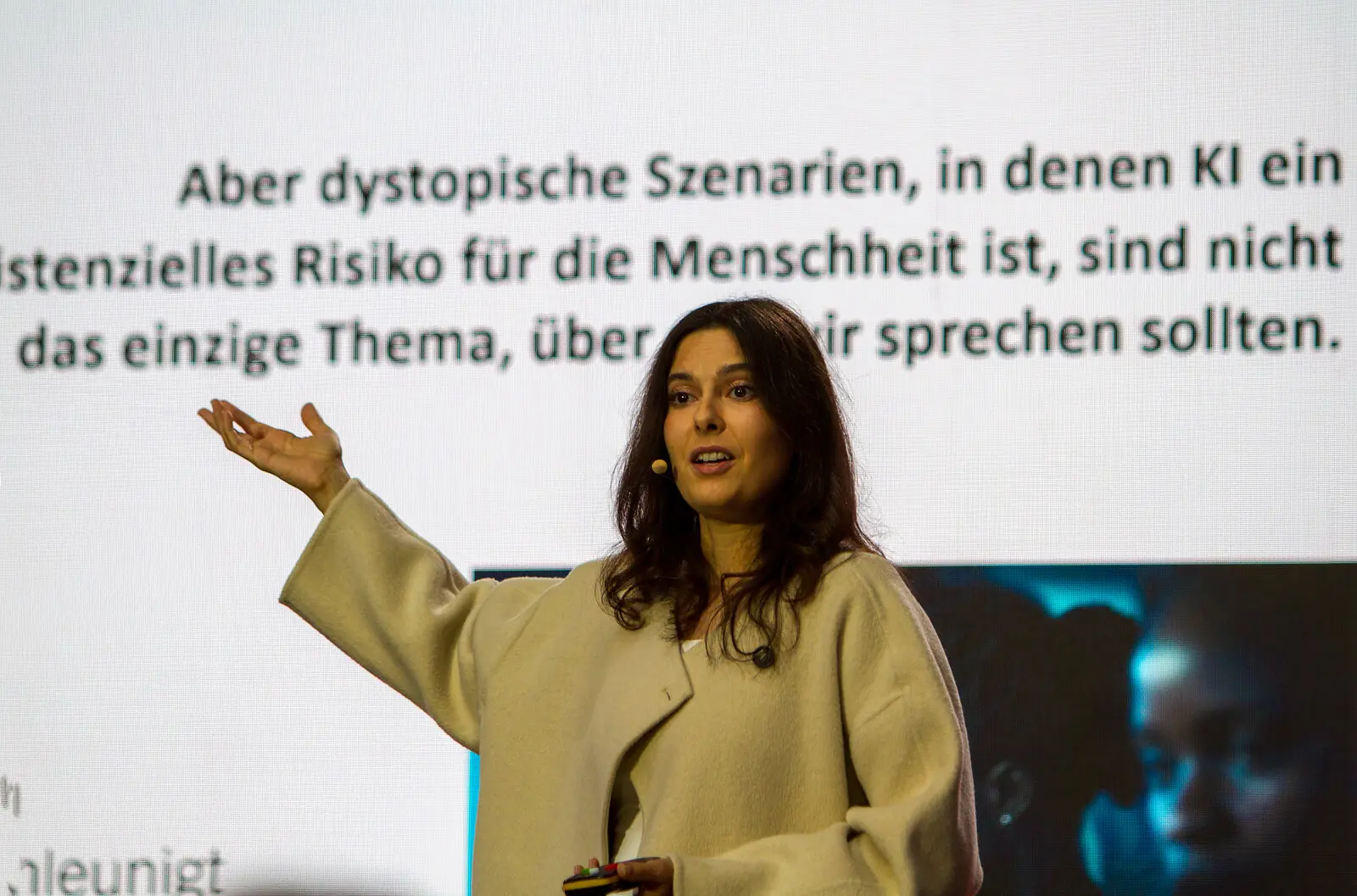 dunkelhaarige Dame mit beigen Blazer und Mikrofon zeigt mit dem rechten Arm auf die Folie und spricht zum Publikum. Auf der Folie ein Bild und der Schriftzug "Aber dystopische Szenarien, in denen KI ein stenzielles Risiko für die Menschheit ist, sind nocht das einzige Thema über wir sprechen sollten.
