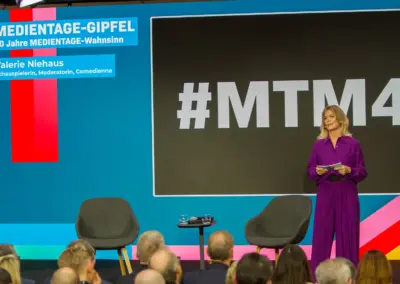 Medientage Bühne Blau - Blauer Hintergrund großes graues Rechteck mit #MTM4 links der Titel in blau weiß MEDIENTAGE-GIPFEL 40 Jahre MEDIENTAGE-WAHNSIINN - darunter der Titel in Blau Weiß Valerie Niehaus Schauspielerin, Moderatorin, Comedienne- zwei leere graue Stühel ein Stehtisch und rechts daneben blonde Frau Valerie Niehaus in Lila Hosenanzug stehend und sprechend. Vordergrund die Hinterköpfe der Zuschauer.