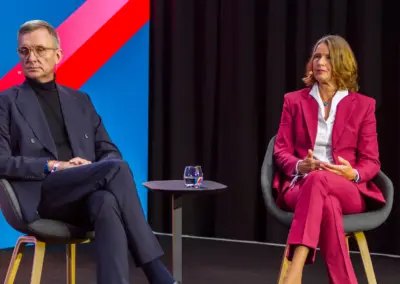 Panel Hintergrund rechts schwarzer Vorhang links blau und pink-roter Streifen . Links im Anzug sitzend Jochen Wegner dazwischen ein Stehtisch mit einem Glas Wasser, rechts sitzend in pink Kostüm und weißer Bluse Prof. Dr. Alexandra Borchardt.