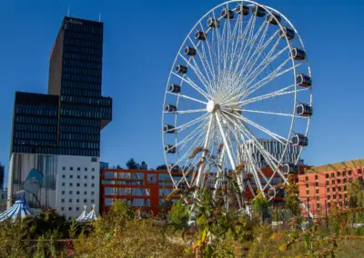 weißes Riesenrad mit Sommerflieder und Garten im Vordergrund. Dahinter rote Hochhäuser und das granitfarbige Bürogebäude und unterhalb die Zirkuszelte. Kunst an der Hauswand mit einem gezeichneten Riesenspiegel.