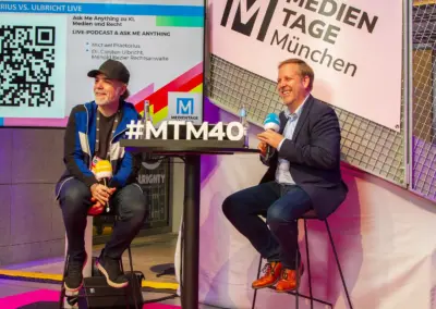 mtm 40 stehtisch links und rechts sitzen zwei männer dahinter die Wendeltreppe des Gebäudes im Rücken die Präsentation des Vortrags und Logo #mtm40, pink schwarz weiße Deko des Raumes.
