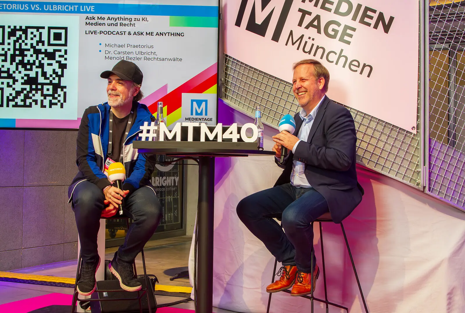 mtm 40 stehtisch links und rechts sitzen zwei männer dahinter die Wendeltreppe des Gebäudes im Rücken die Präsentation des Vortrags und Logo #mtm40, pink schwarz weiße Deko des Raumes.