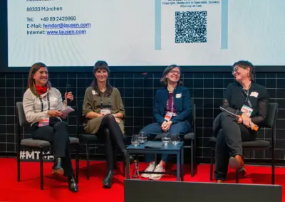 roter Fußboden, schwarze Fliesen dahinter die Präsentation von lausen.com - von links nach rechts sitzend und lachend im Gespräch Dr. Ursula Feindor-Schmidt (Kanzlei Lausen), Dr. Armelle Grandjean und Christina von Gemmingen-Hornberg (GEMA), Moderation Anja Braune. 22. Oktober 2026.