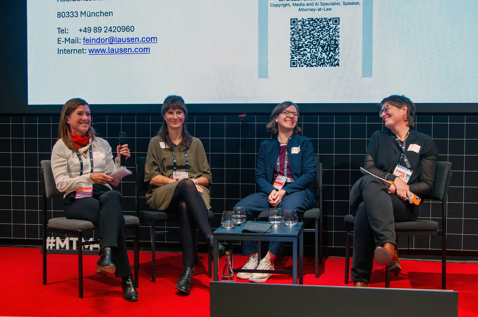 roter Fußboden, schwarze Fliesen dahinter die Präsentation von lausen.com - von links nach rechts sitzend und lachend im Gespräch Dr. Ursula Feindor-Schmidt (Kanzlei Lausen), Dr. Armelle Grandjean und Christina von Gemmingen-Hornberg (GEMA), Moderation Anja Braune. 22. Oktober 2026.