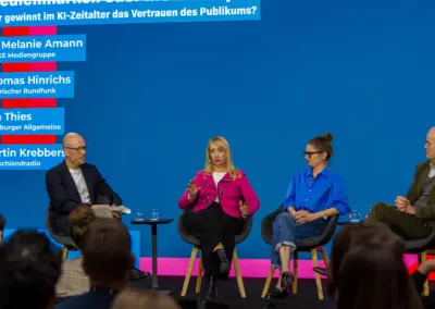 auf der Bühne mit blauem Hintergrund die Teilnehmer sitzend im Gespräch. Dr. Melanie Amann (FUNKE Mediengruppe), Thomas Hinrichs (Bayerischer Rundfunk), Lea Thies und Martin Krebbers (Deutschlandradio), 23. Oktober 2026.