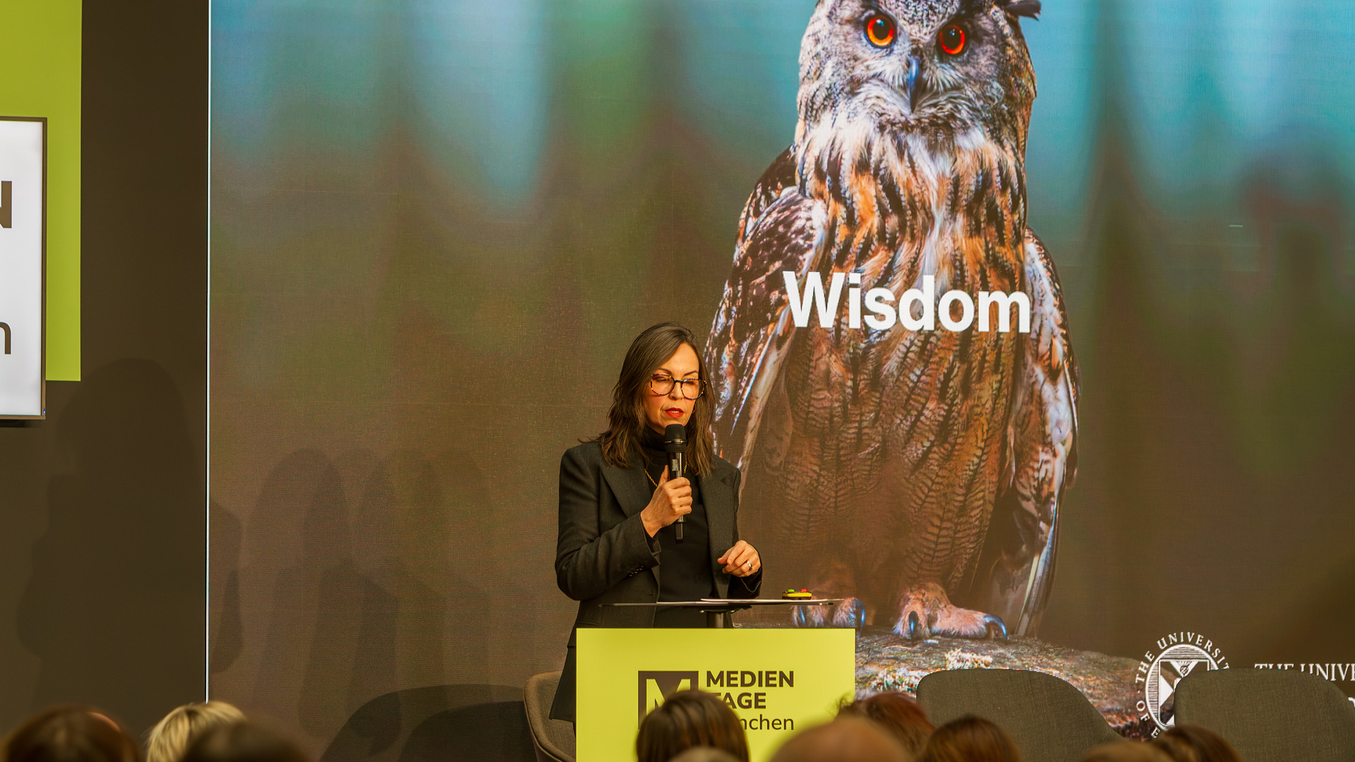 im Hintergrund eine große Eule mit der Aufschrift Wisdom in weißer Schrift - am Rednerpult redend in das Mikrofon. Prof. Shannon Vallor, in schwarzem Kostüm.