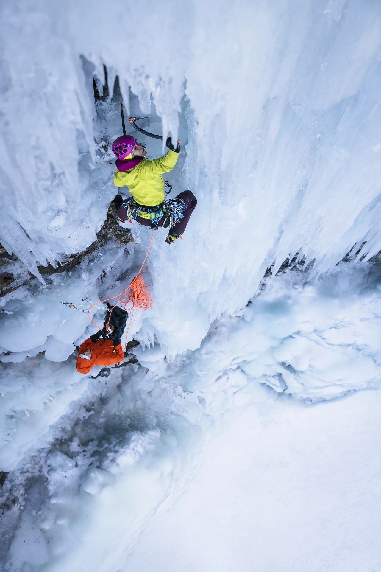 Eine Kletterin in gelber Jacke und violettem Helm steigt mit Eisgeraeten eine steile Eiswand hinauf. Weiter unten sichert eine zweite Person in oranger Jacke am orangefarbenen Seil.