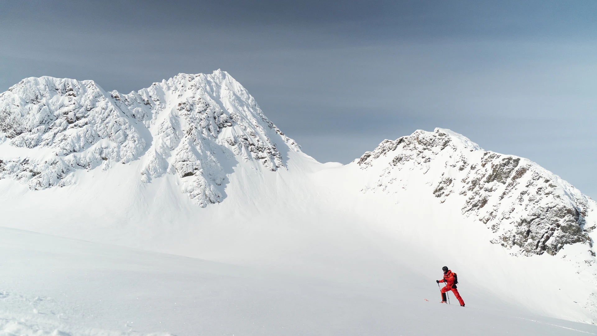 Ein einzelner Skitourengeher in roter Kleidung steigt ueber ein weites Schneefeld, dahinter zwei verschneite Felsgipfel unter grauem Himmel.