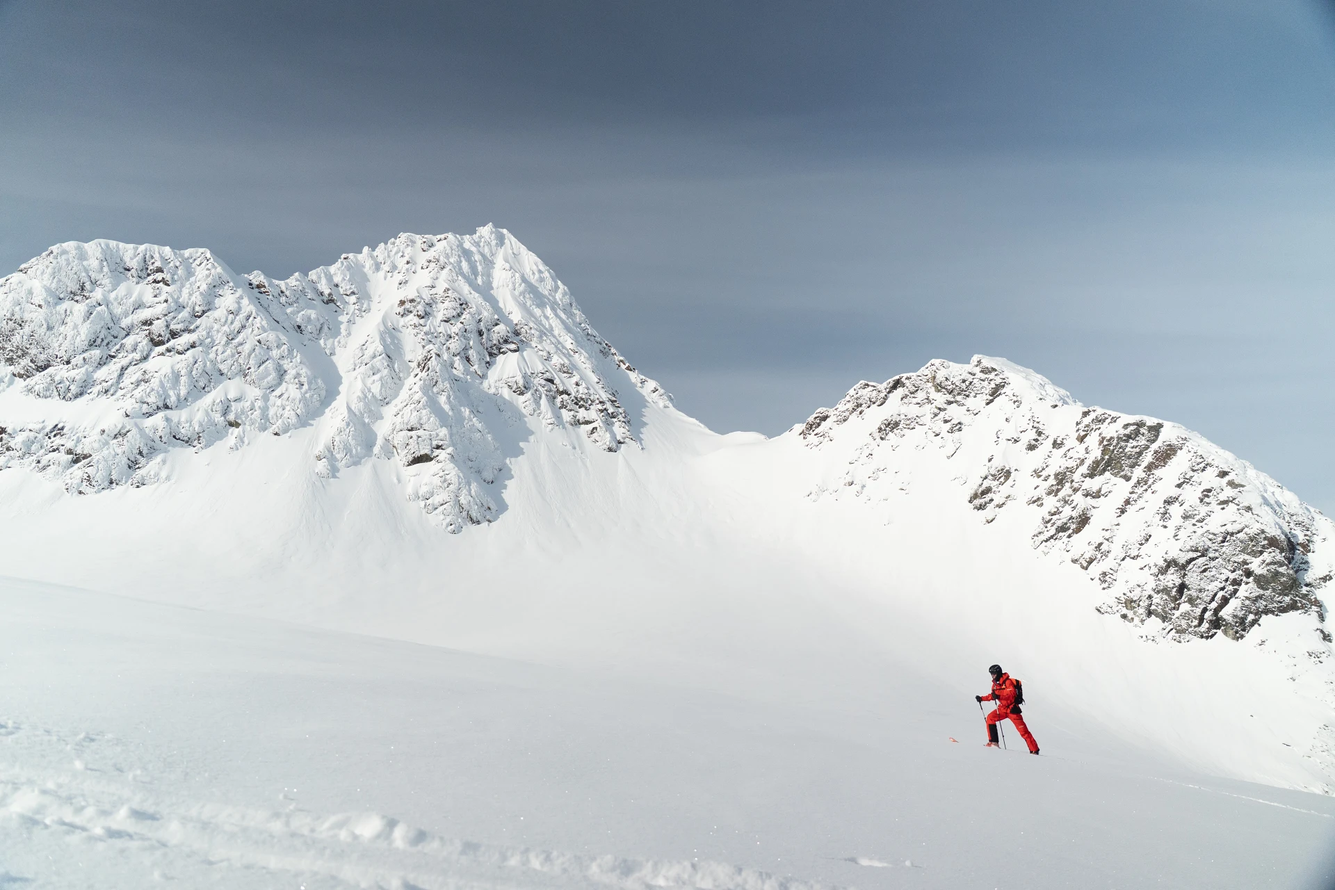 Ein einzelner Skitourengeher in roter Kleidung steigt ueber ein weites Schneefeld, dahinter zwei verschneite Felsgipfel unter grauem Himmel.