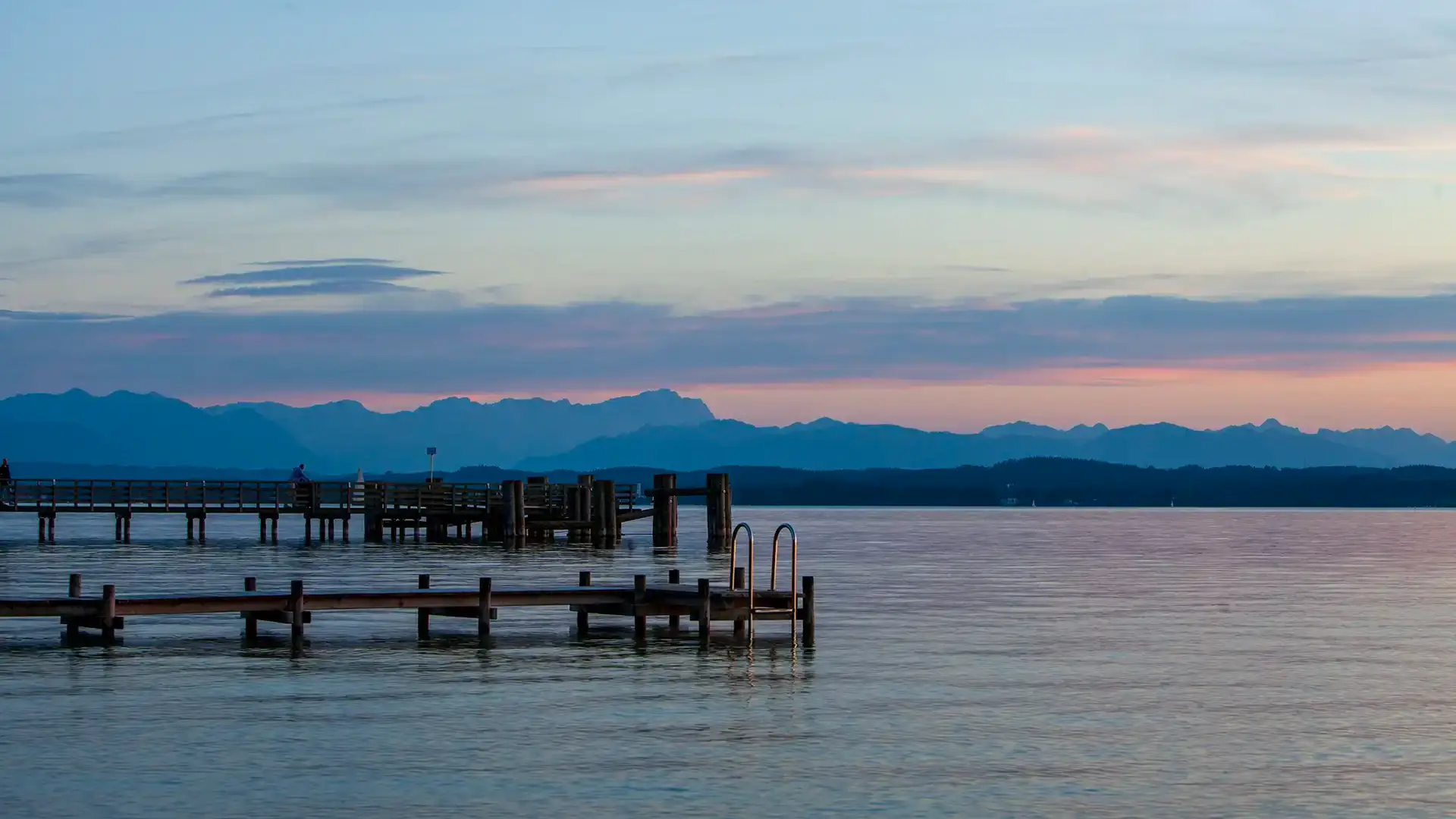 Abendstimmung über dem Starnberger See. Im Vordergrund das Wasser des Sees in rot blauer Färbung. Zwei Holzstege ins Wasser. Im Hintergrund die Alpenkette in rotem Licht und leichte Wolken am Himmel.