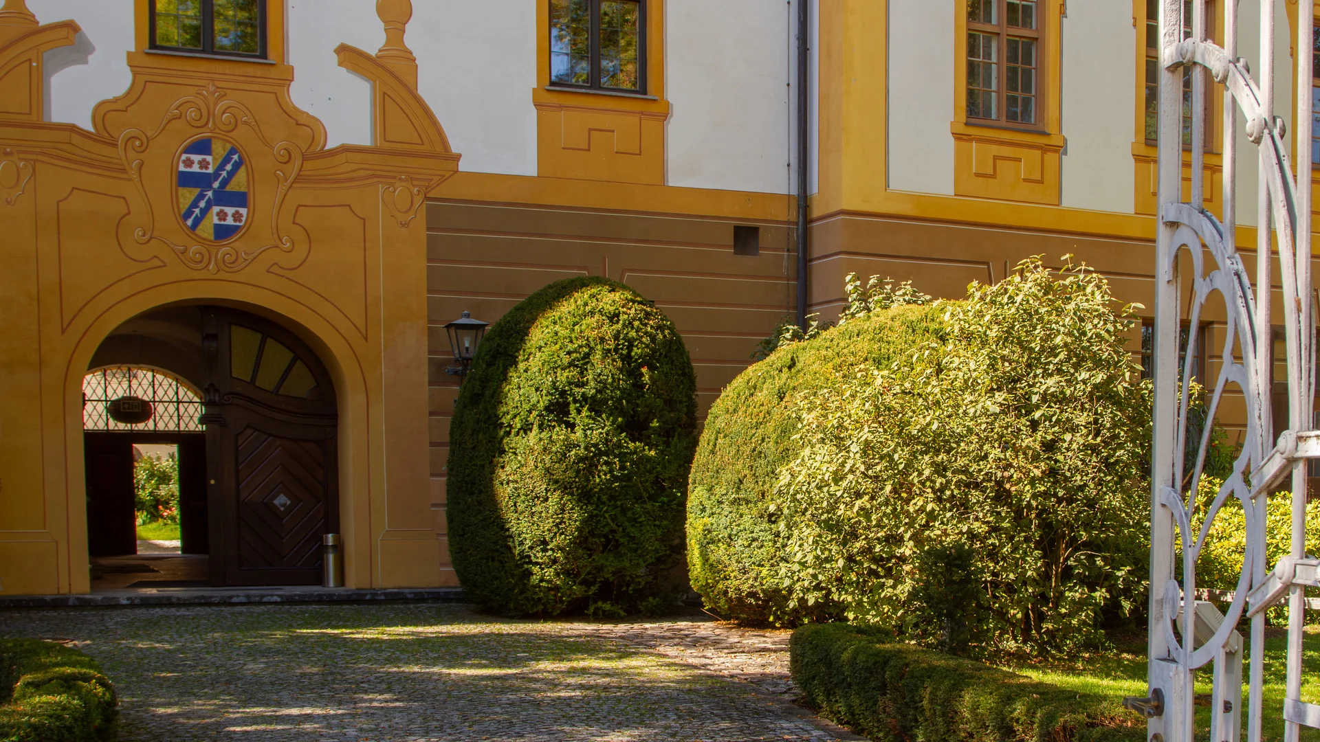 Barockes Torhaus mit Wappen ueber dem Rundbogen, davor ein gepflasterter Weg und geschnittene Buchskugeln; rechts ein weisses schmiedeeisernes Tor.