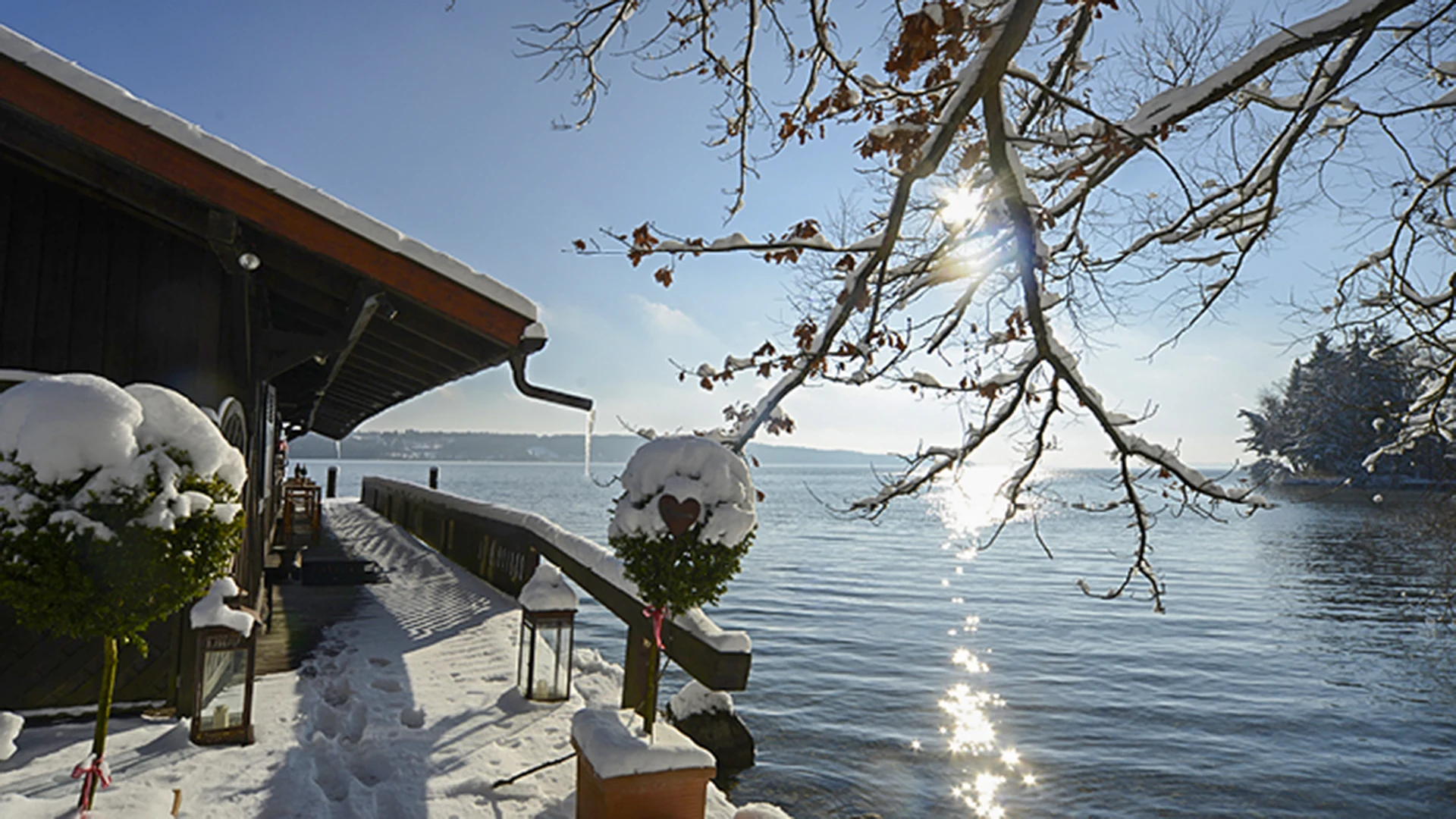 Ein verschneiter Holzsteg fuehrt an einem dunklen Bootshaus entlang zum Ufer eines Sees. Die tief stehende Sonne bricht als Stern durch schneebedeckte Eichenaeste, auf dem Wasser glitzert ihr Licht. Vorn steht ein Buchsbaumstaemmchen mit Schneehaube, in das ein Herz gesteckt ist, am Stamm haengt eine rosa Schleife. Daneben eine Laterne im Schnee.